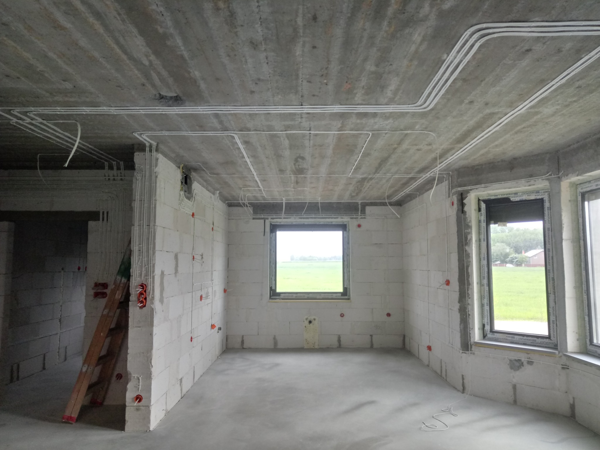 Wnętrze w stanie surowym: białe ściany z betonu komórkowego, widoczne przewody elektryczne na suficie i ścianach, okna z widokiem na zieleń, drabina oparta o ścianę.