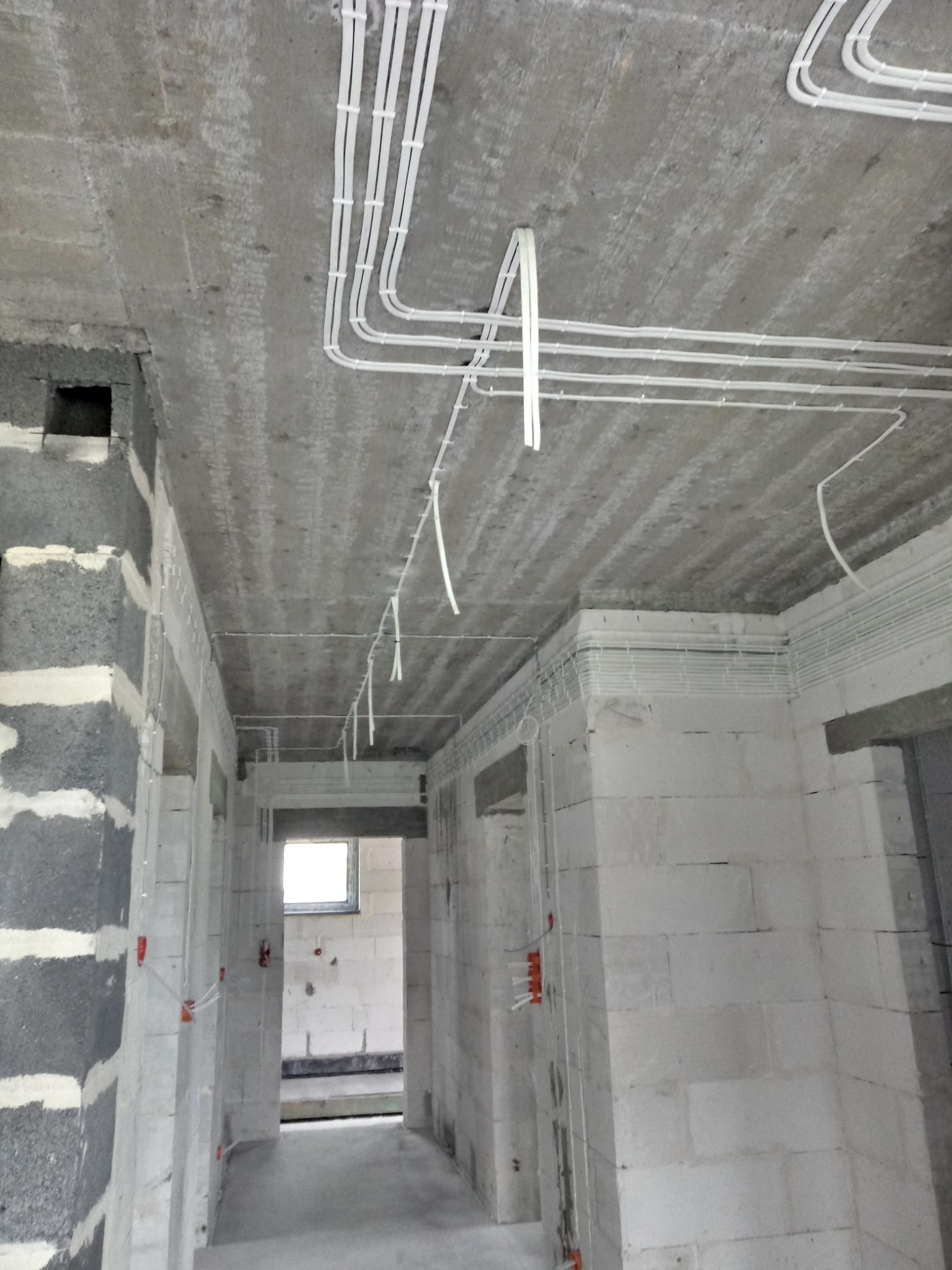 Białe kable elektryczne ułożone na suficie z surowego betonu w budynku w trakcie budowy. Widoczne ściany z białych bloczków i otwory drzwiowe.