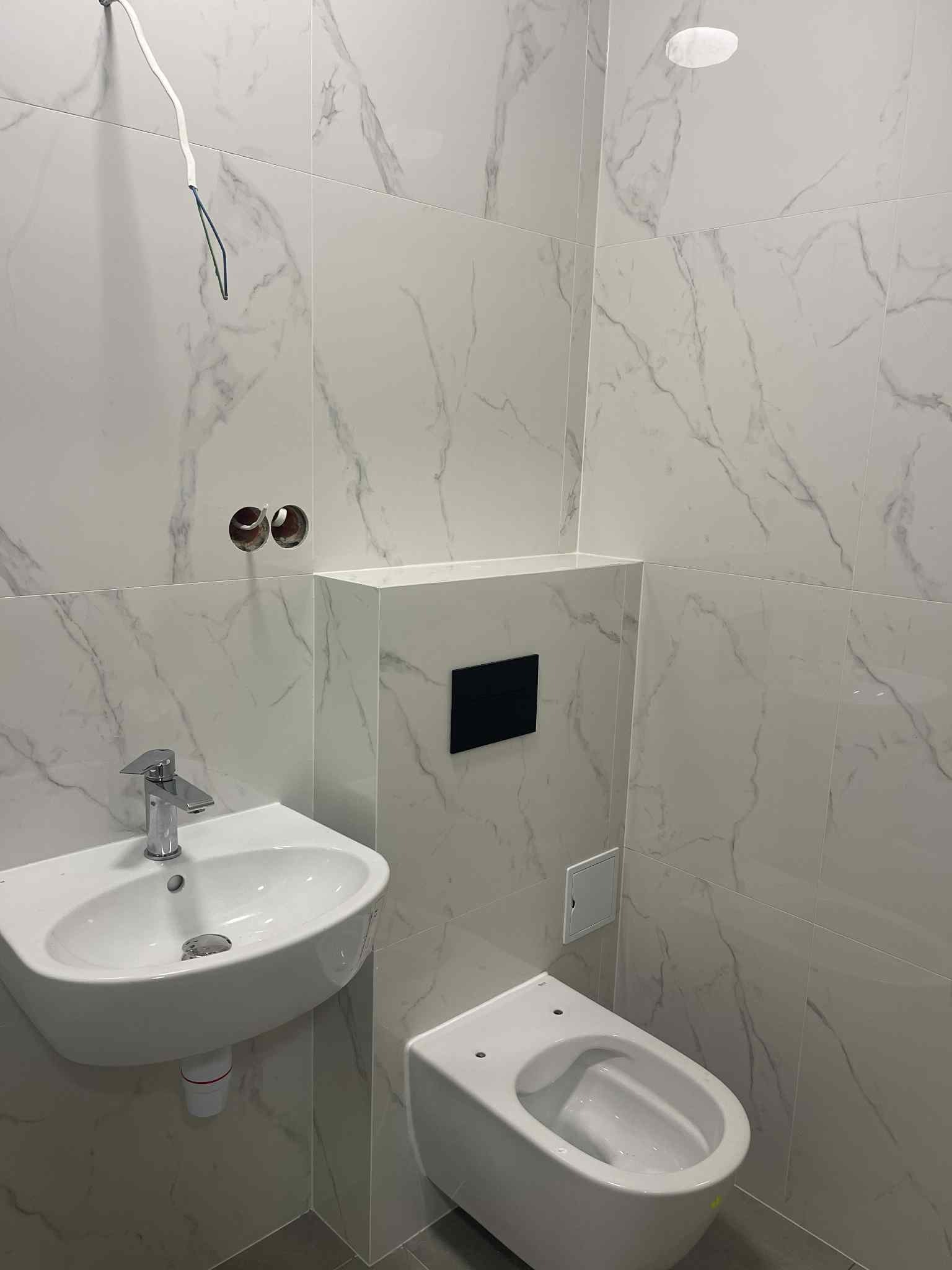 Wykończona łazienka z marmurowymi płytkami, wiszącą toaletą i umywalką. Widoczne niedokończone podłączenia elektryczne. Minimalistyczny design.
