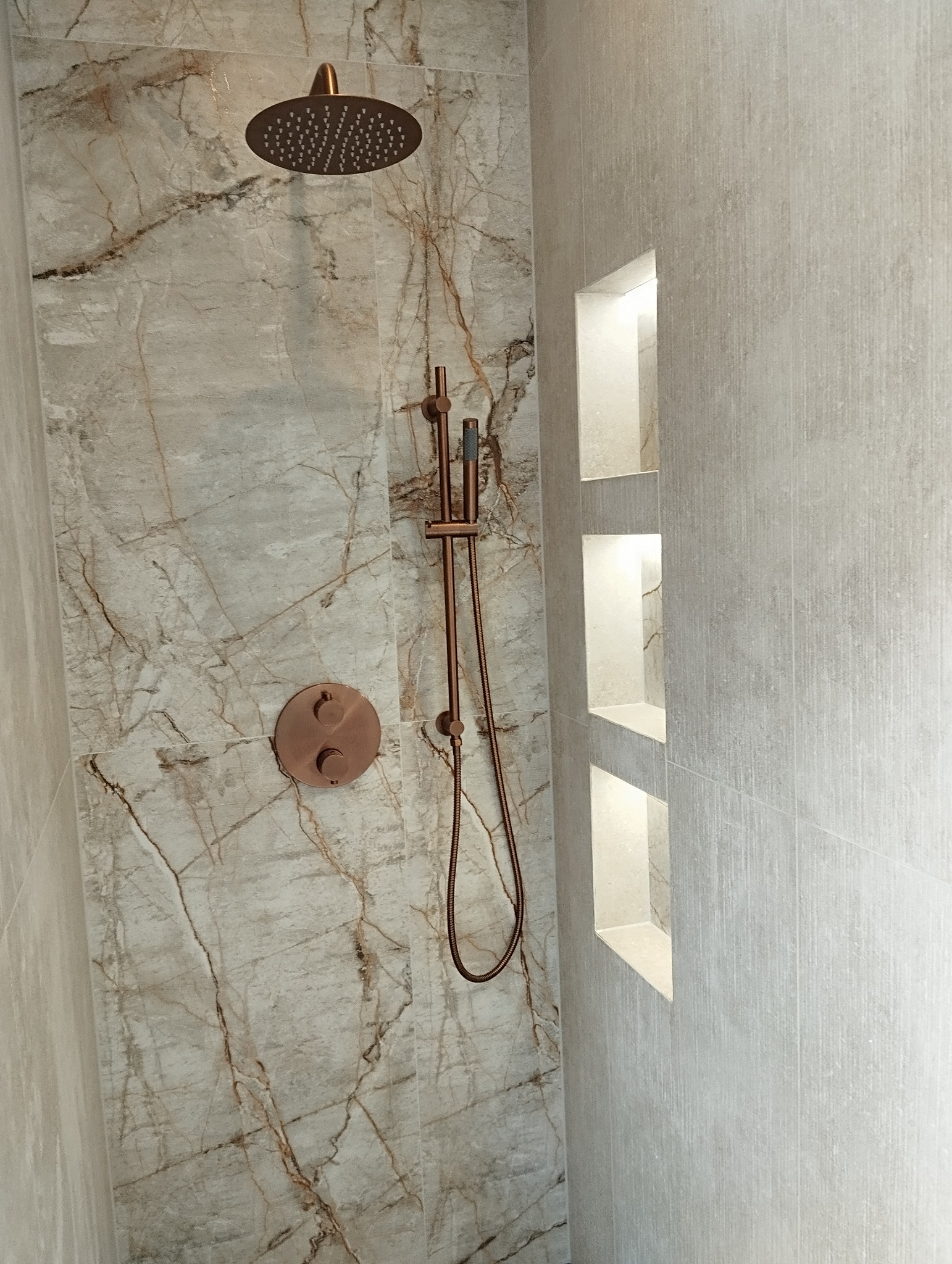 Wykończona kabina prysznicowa z marmurowymi płytkami i miedzianą armaturą. W ścianie wnęki z oświetleniem. Minimalistyczny design.