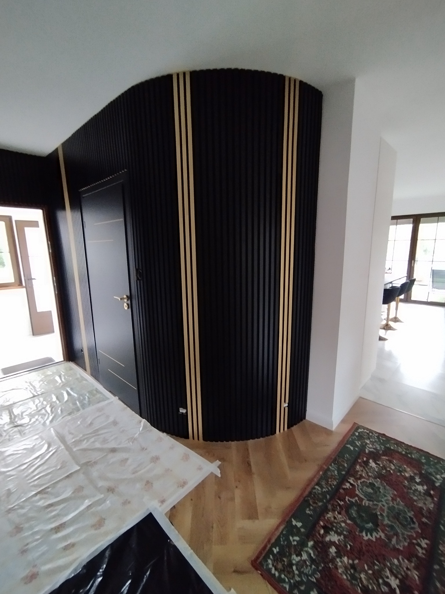 Wykończona ściana z czarnych paneli pionowych z jasnymi paskami, obok czarne drzwi z poziomymi zdobieniami. Podłoga zabezpieczona folią malarską.