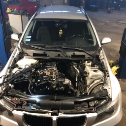 Tomasz Czernik - Srebrny samochód na podnośniku w warsztacie z otwartą maską, widoczne elementy silnika i narzędzia, w tle inne pojazdy i wyposażenie warsztatowe.