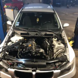 Srebrny samochód na podnośniku w warsztacie z otwartą maską, widoczne elementy silnika i narzędzia, w tle inne pojazdy i wyposażenie warsztatowe.