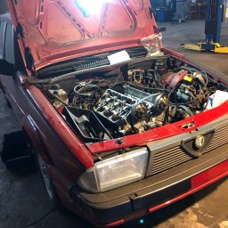 Tomasz Czernik - Czerwony samochód Alfa Romeo z otwartą maską w warsztacie, widoczne elementy silnika, w trakcie naprawy, zarys wnętrza warsztatu z podnośnikiem.