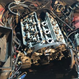 Tomasz Czernik - Otwarty silnik samochodowy w trakcie naprawy, widoczne głowice cylindrów, pasek rozrządu i liczne przewody, czerwona karoseria w tle.