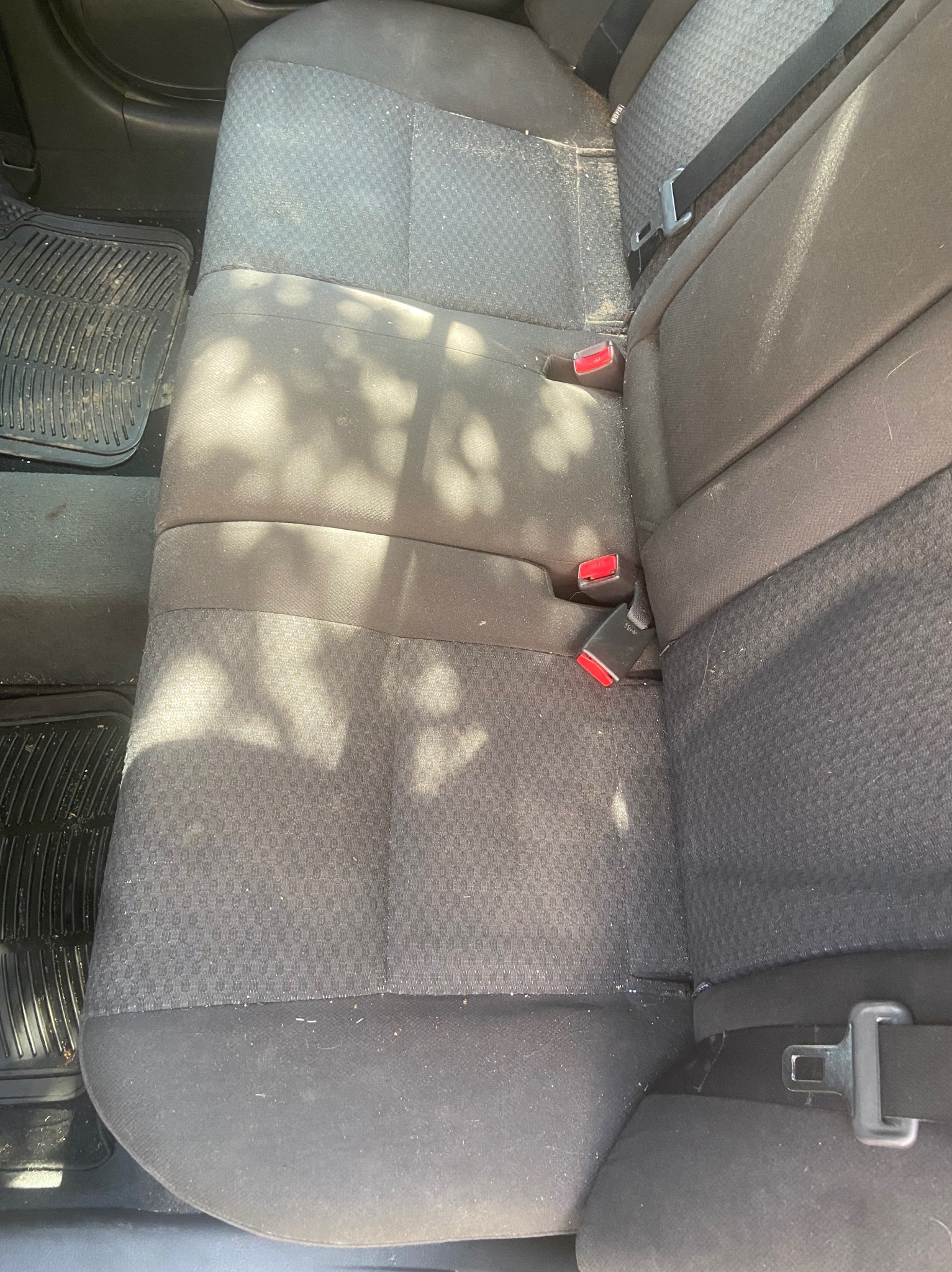 Widok z góry na tylną kanapę samochodu z widocznymi plamami, okruchami i sierścią zwierząt, sugerującymi potrzebę czyszczenia. Widoczny fragment gumowej wycieraczki pod nogami.
