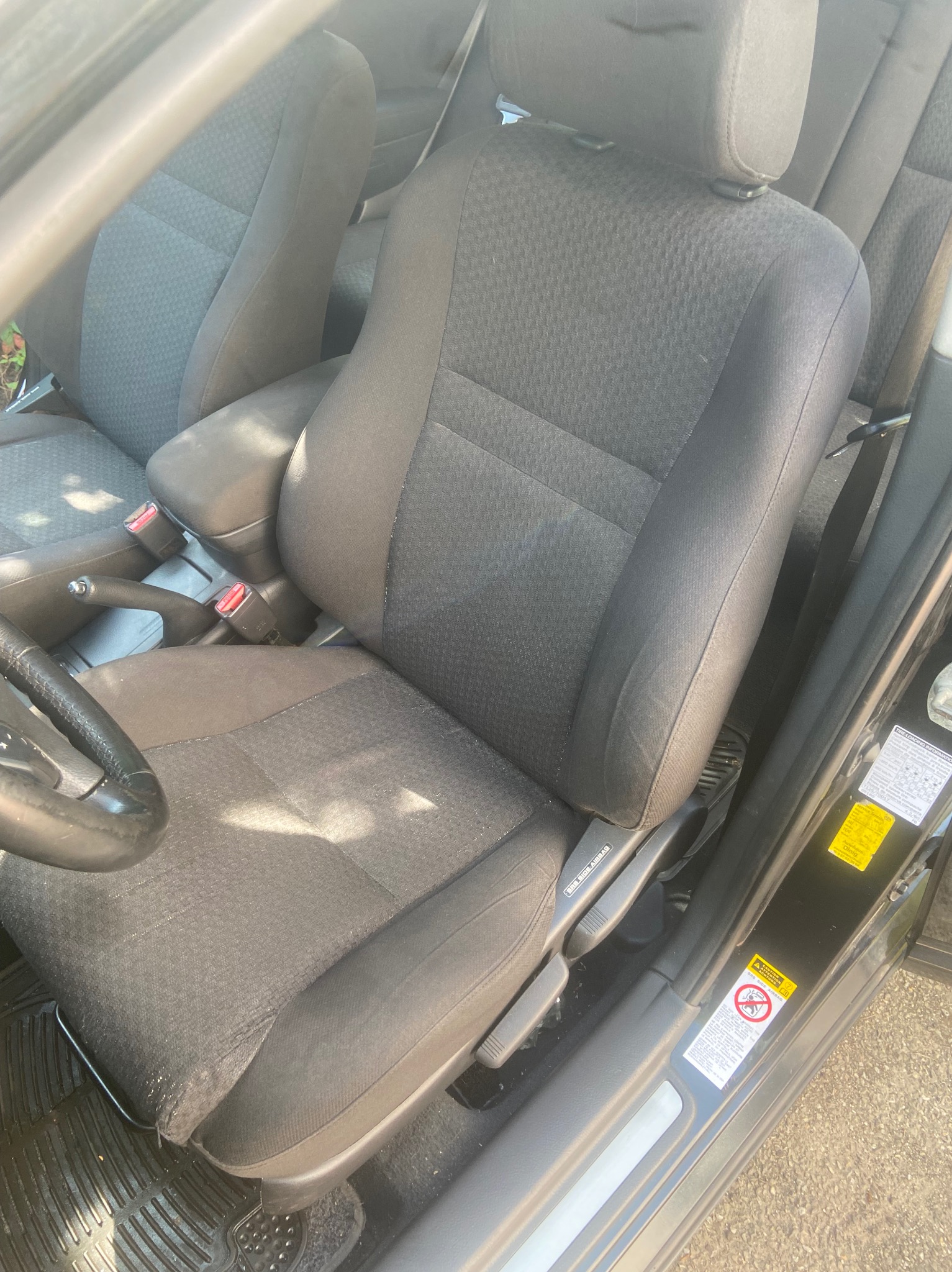 Wnętrze samochodu z widocznym czarnym fotelem kierowcy z regulacją, gumowa wycieraczka pod nogami oraz naklejki informacyjne na progu.