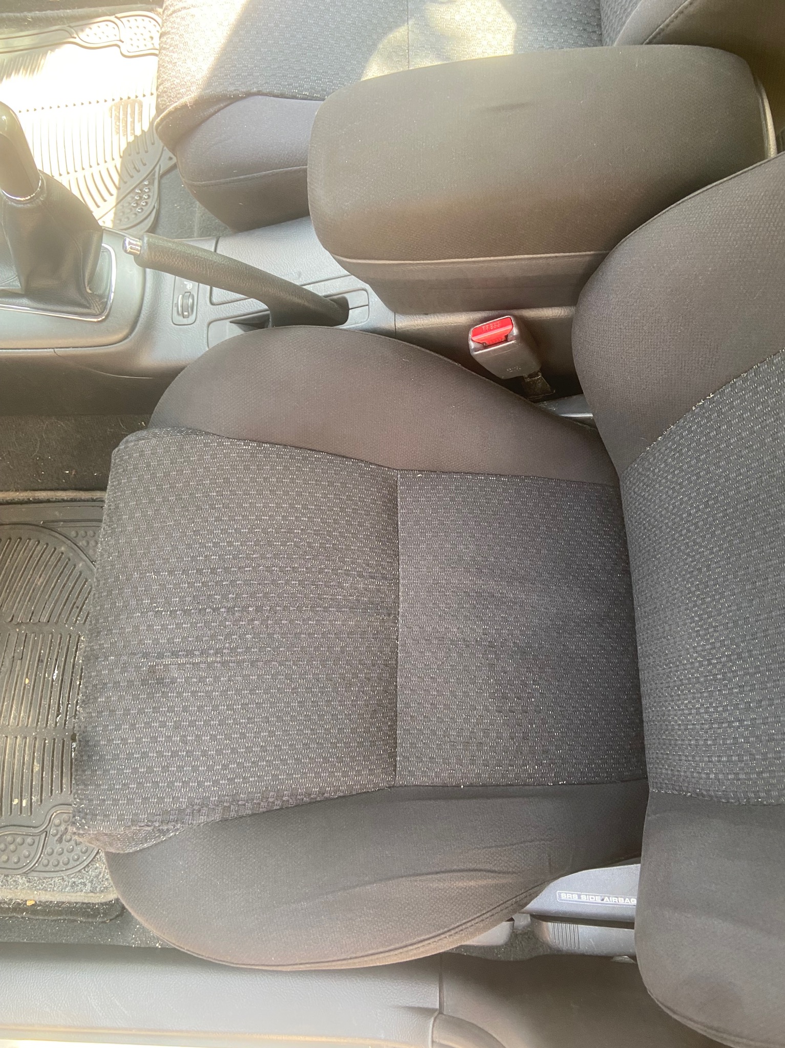 Wnętrze samochodu z widocznym fotelem kierowcy z czarną tapicerką, podłogą z gumową wycieraczką, dźwignią zmiany biegów, hamulcem ręcznym i podłokietnikiem, przed praniem tapicerki.