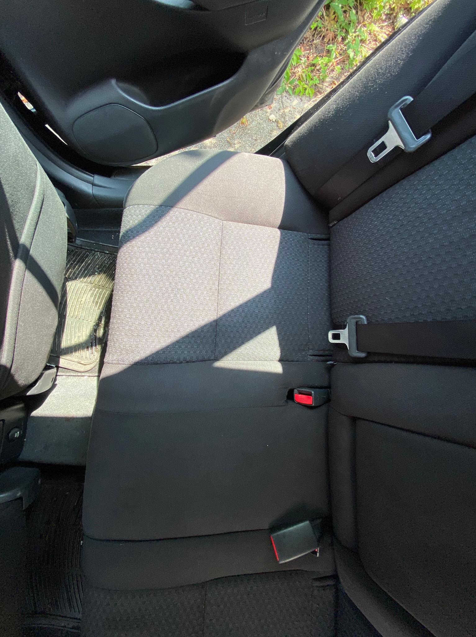 Wnętrze samochodu z widoczną tylną kanapą, pasami bezpieczeństwa i gumowymi dywanikami, częściowo oświetlone przez słońce wpadające przez otwarte drzwi.