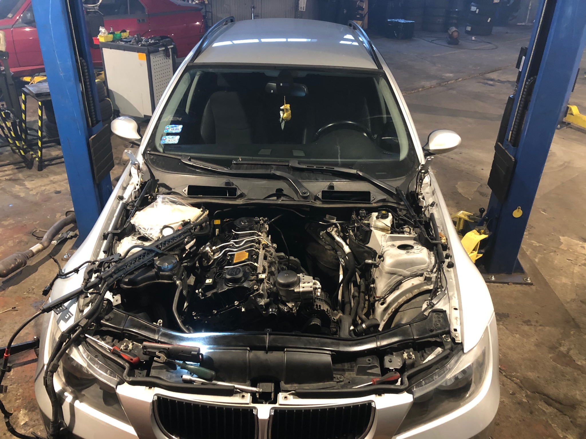 Srebrny samochód na podnośniku w warsztacie z otwartą maską, widoczne elementy silnika i narzędzia, w tle inne pojazdy i wyposażenie warsztatowe.