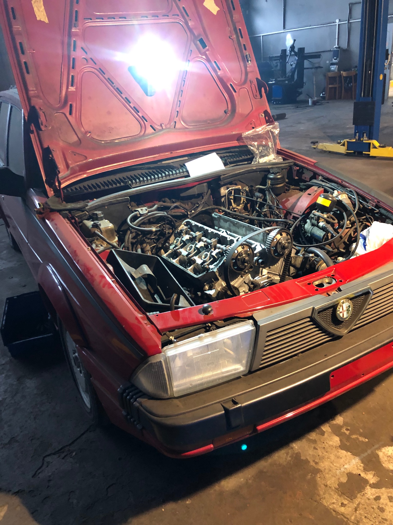 Czerwony samochód Alfa Romeo z otwartą maską w warsztacie, widoczne elementy silnika, w trakcie naprawy, zarys wnętrza warsztatu z podnośnikiem.