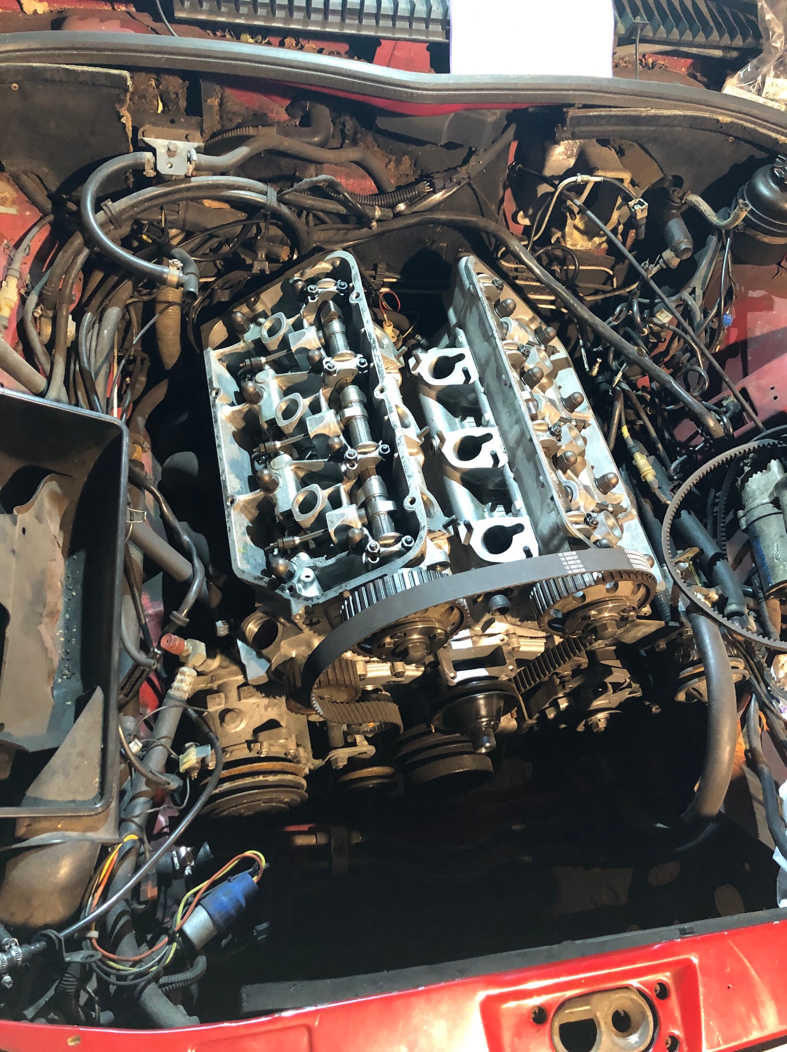 Otwarty silnik samochodowy w trakcie naprawy, widoczne głowice cylindrów, pasek rozrządu i liczne przewody, czerwona karoseria w tle.