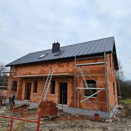 Wojciech Podborski - Dom w trakcie budowy z cegły, z zamontowanym czarnym dachem z blachy na rąbek stojący, rusztowania przy ścianach i stertą cegieł na palecie przed budynkiem. Widoczny pracownik w oddali.
