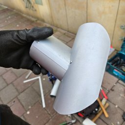 Wojciech Podborski - Blaszany element rynny w rękawicy roboczej, widoczne nity, narzędzia i fragment elewacji budynku w tle.