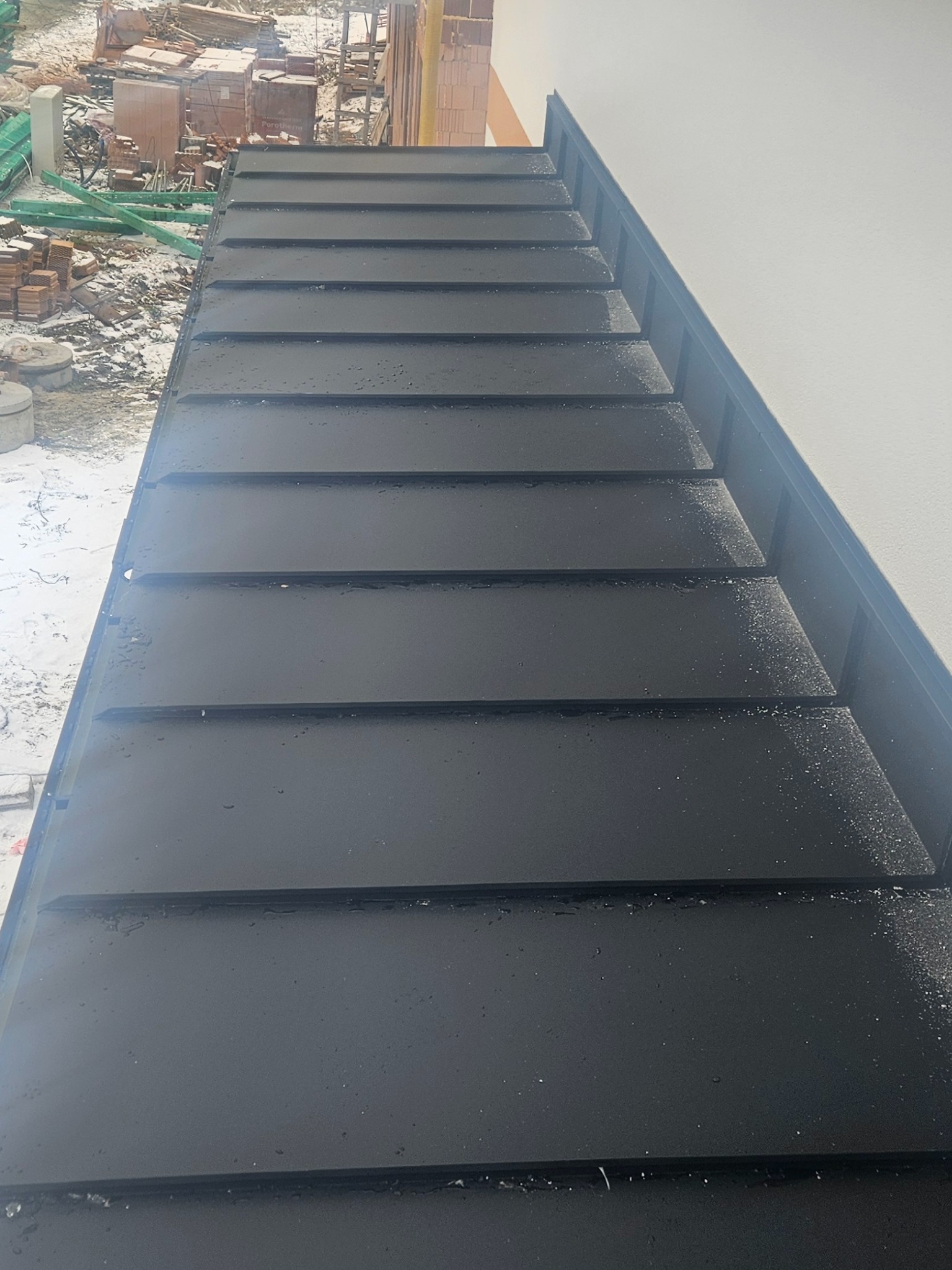 Ciemna, metalowa powierzchnia dachu z widocznymi łączeniami, częściowo pokryta drobnymi kroplami wody lub szronem, z fragmentem białej ściany budynku po prawej stronie i elementami placu budowy w tle.