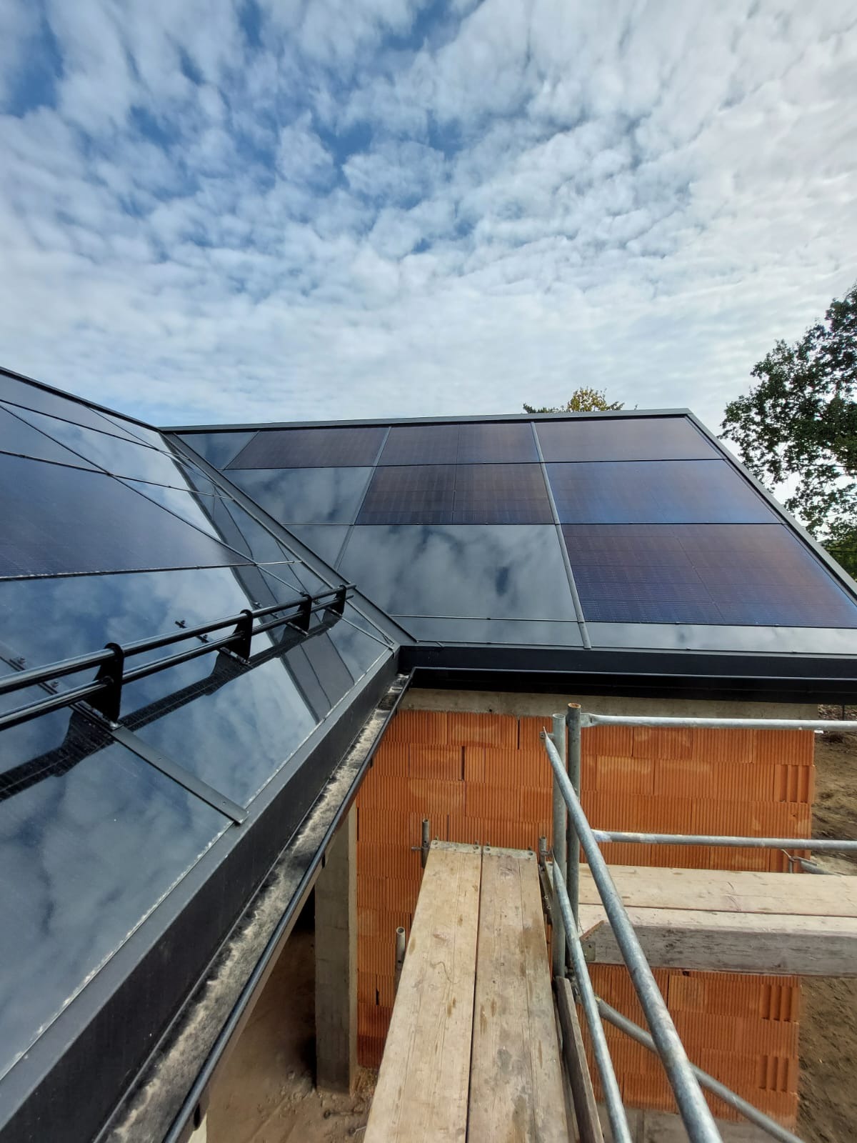 Nowoczesny dach z panelami fotowoltaicznymi w trakcie montażu, widoczna konstrukcja nośna, rusztowanie i fragment muru z czerwonej cegły, niebo z chmurami odbija się w panelach.