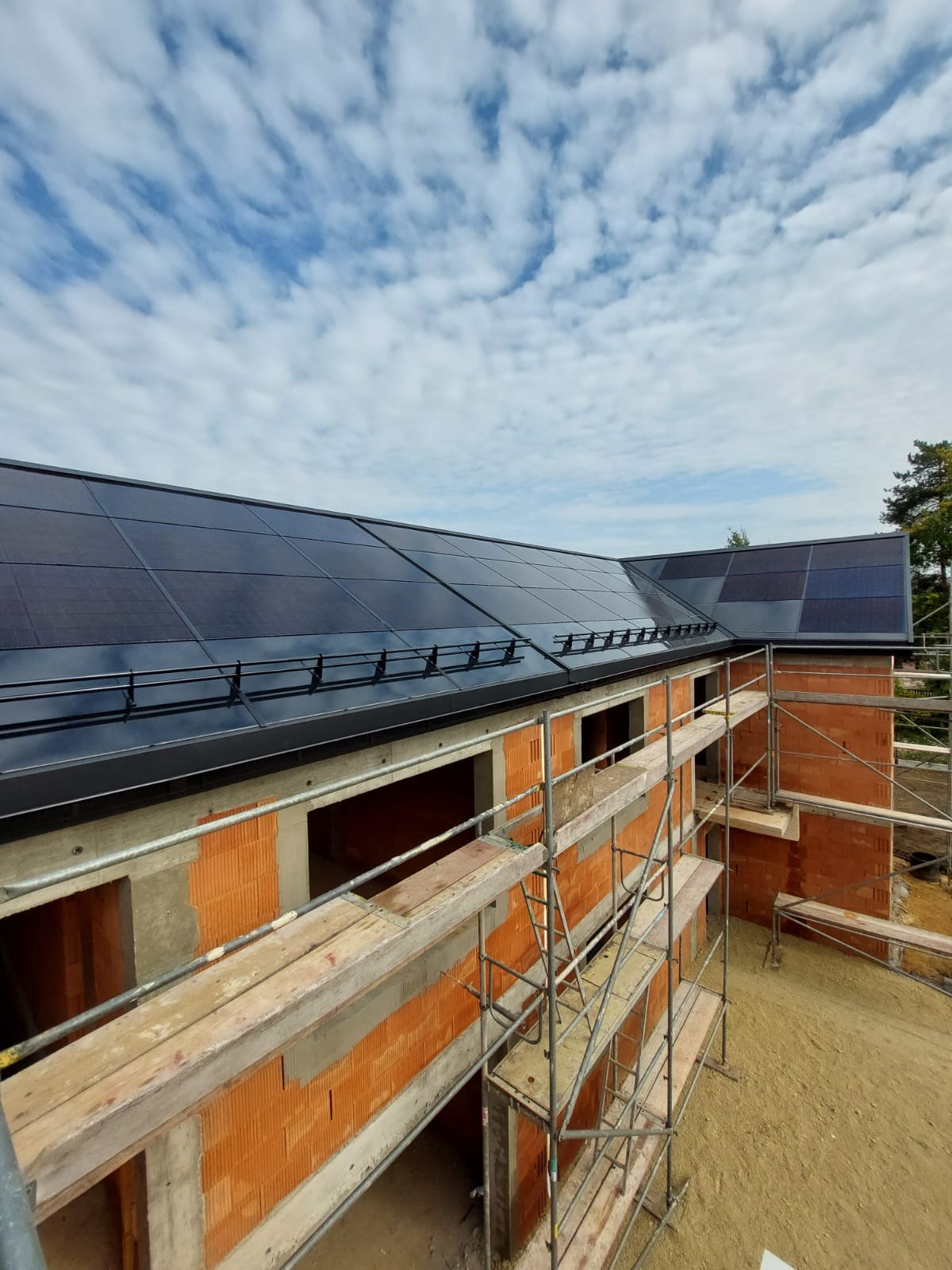 Niedokończony budynek z czerwonej cegły z zamontowanymi panelami słonecznymi na czarnym dachu, widoczne rusztowania i błękitne niebo z chmurami.