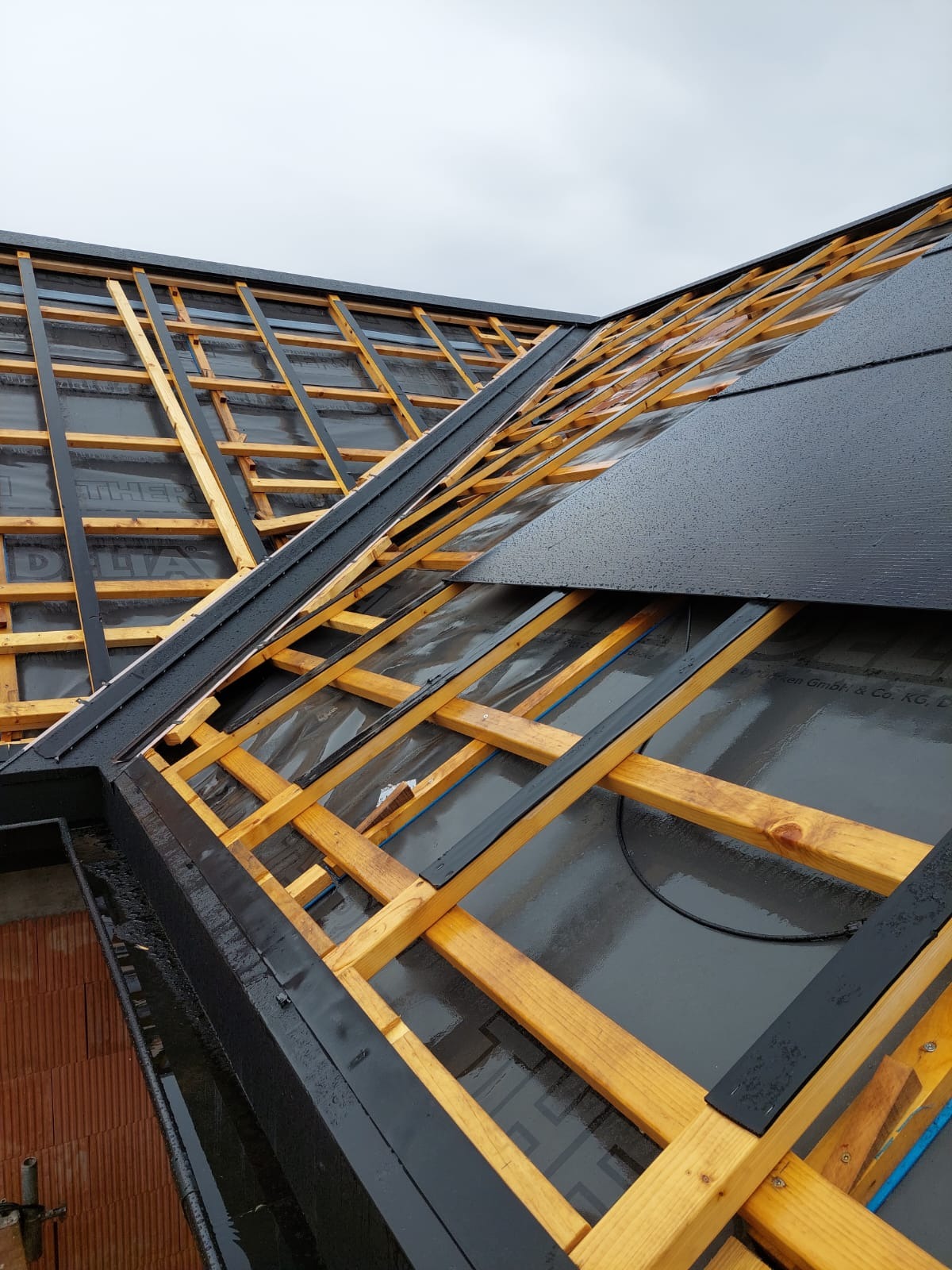 Konstrukcja drewniana dachu z czarną membraną i fragmentem czarnego pokrycia, widoczne krople wody po deszczu.