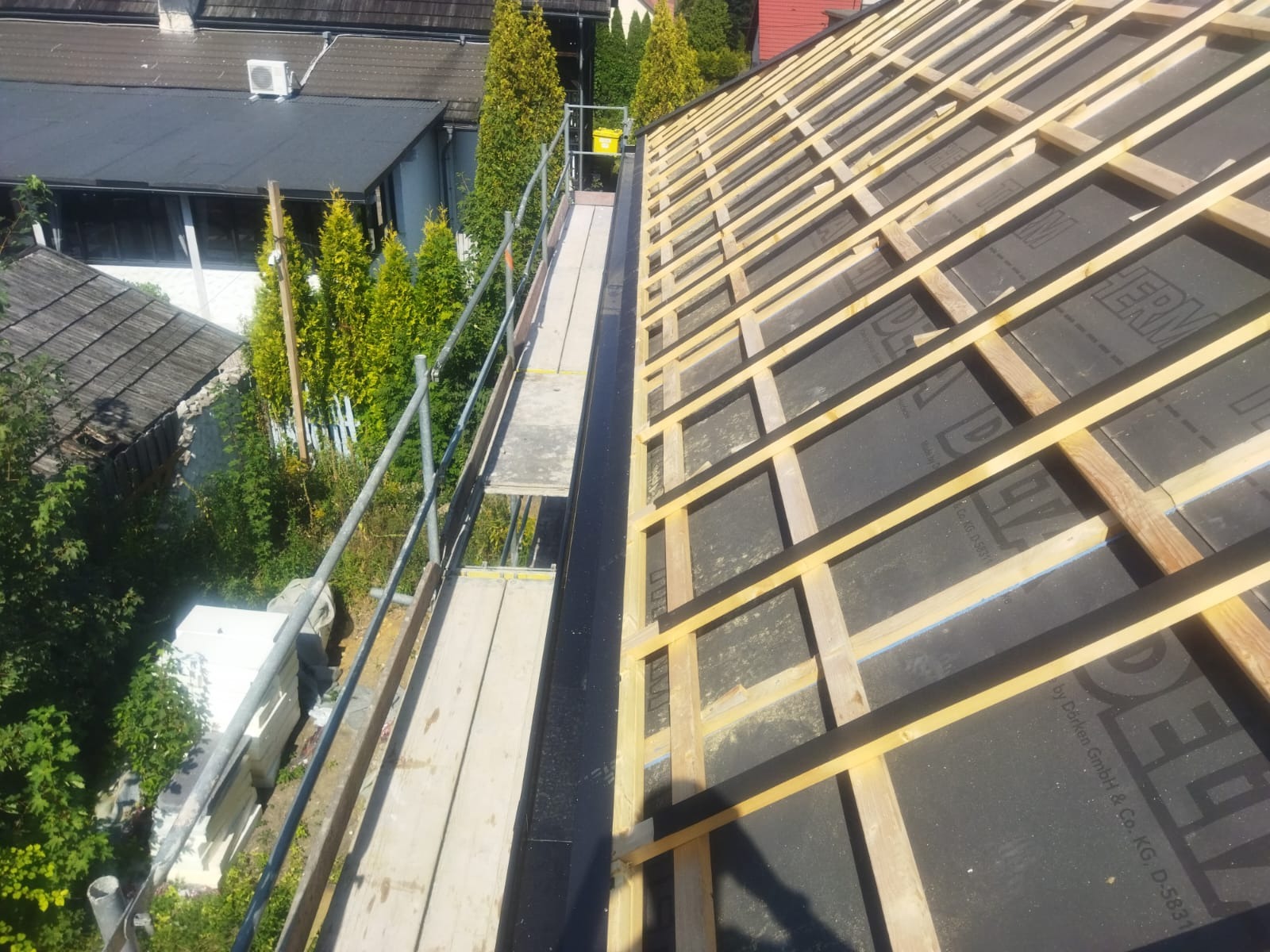 Konstrukcja dachu z łatami i kontrłatami pokryta membraną dachową, widok z rusztowania na tle zieleni i budynków.