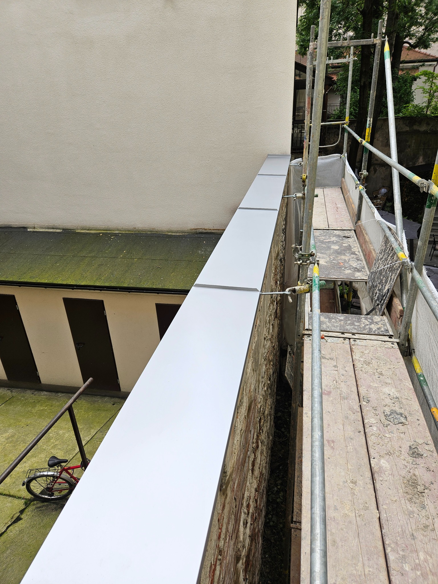 Nowe, jasnoszare obróbki blacharskie na krawędzi dachu, widok z rusztowania, z rowerem opartym o ścianę w tle.