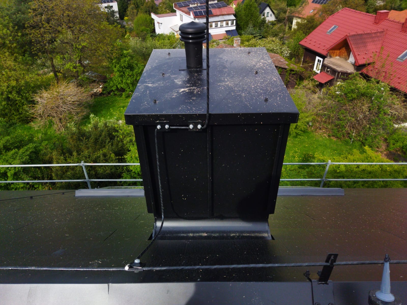 Czarny komin wentylacyjny na dachu pokrytym papą, z widocznymi elementami mocującymi i otoczeniem zieleni oraz zabudowań.