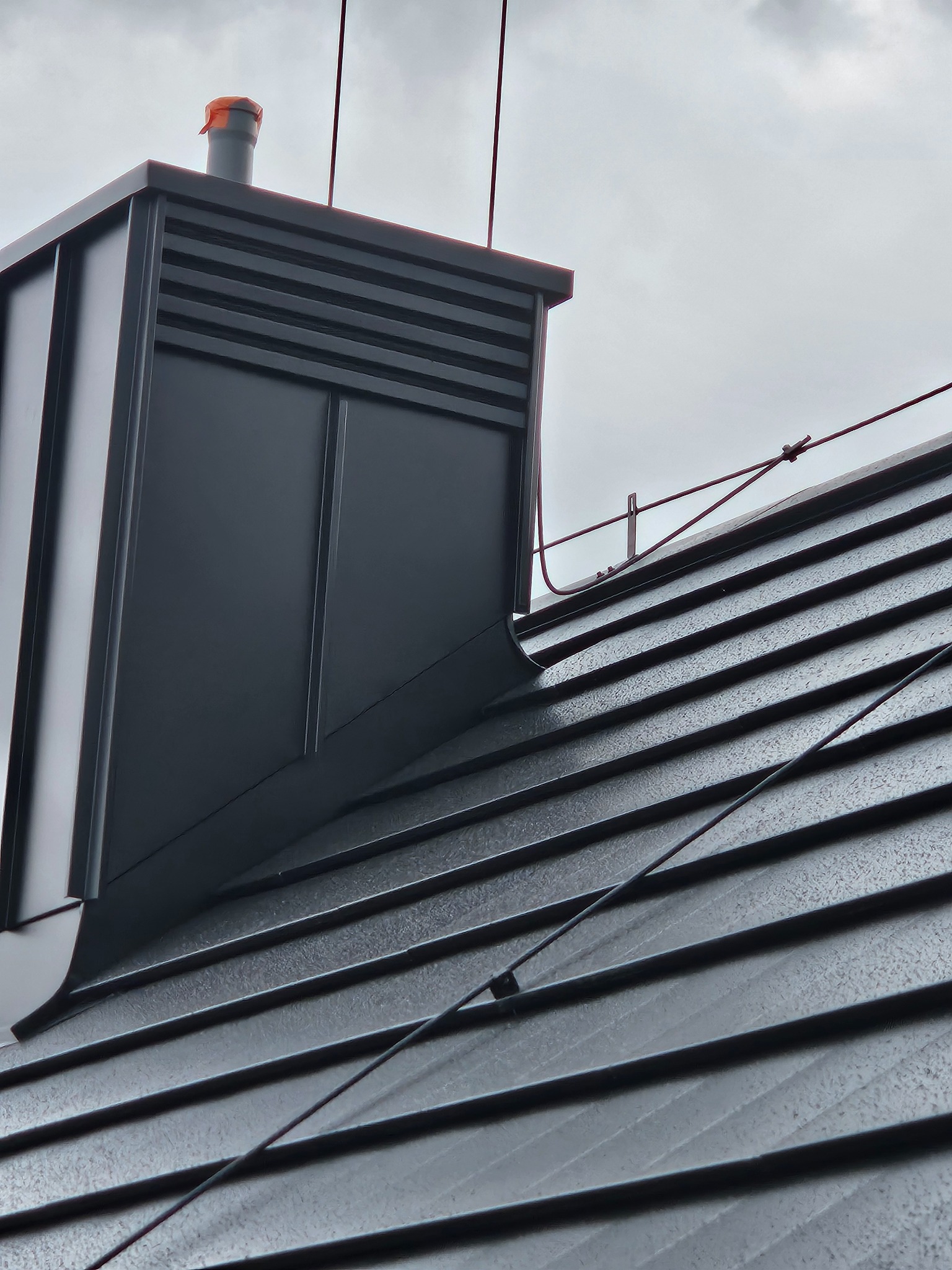 Nowoczesny komin wentylacyjny z czarnej blachy na dachu pokrytym ciemną dachówką ceramiczną, widok z dołu na tle pochmurnego nieba.