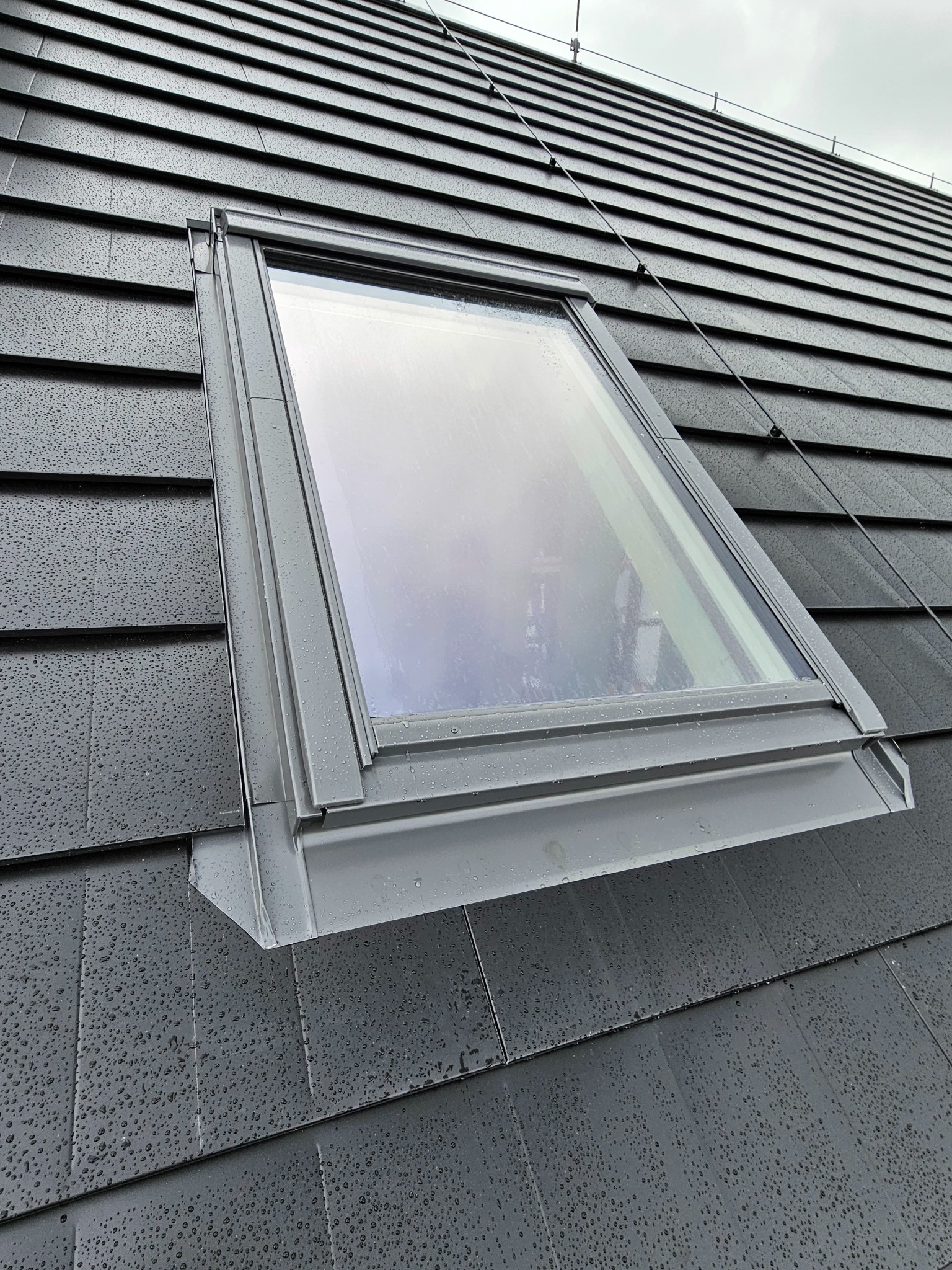 Szare okno dachowe zamontowane w ciemnoszarym dachu pokrytym dachówką, widoczne krople deszczu na powierzchni.