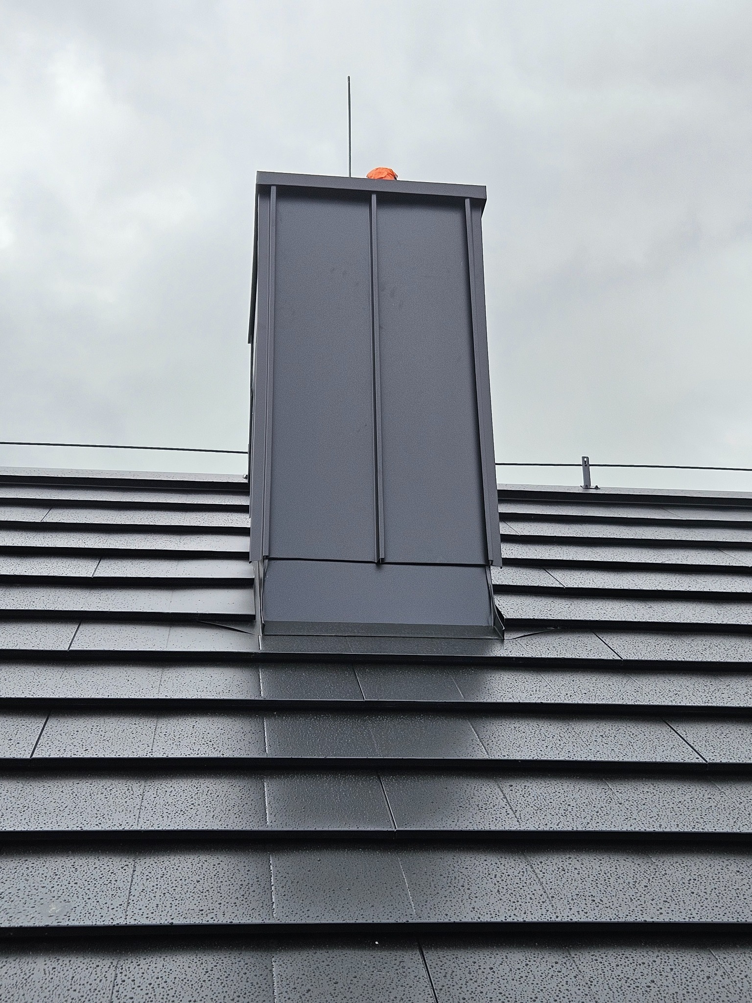 Nowoczesny, grafitowy komin z blachy na dachu pokrytym ciemną dachówką modułową, widoczne krople deszczu, pochmurne niebo.