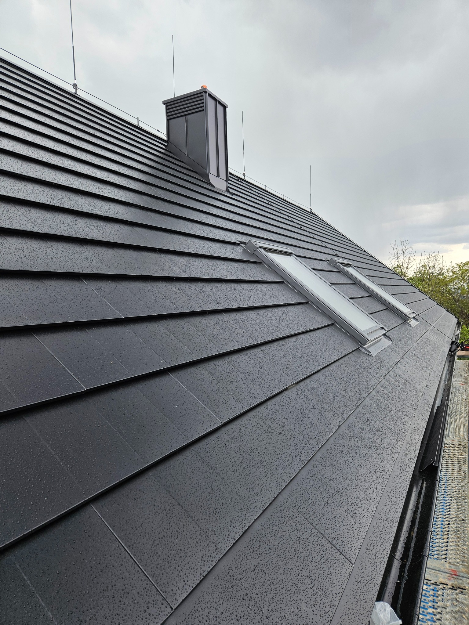Nowoczesny dach pokryty czarną dachówką z dwoma oknami dachowymi i kominem w kolorze grafitowym, widoczne zabezpieczenia odgromowe i fragment rynny.
