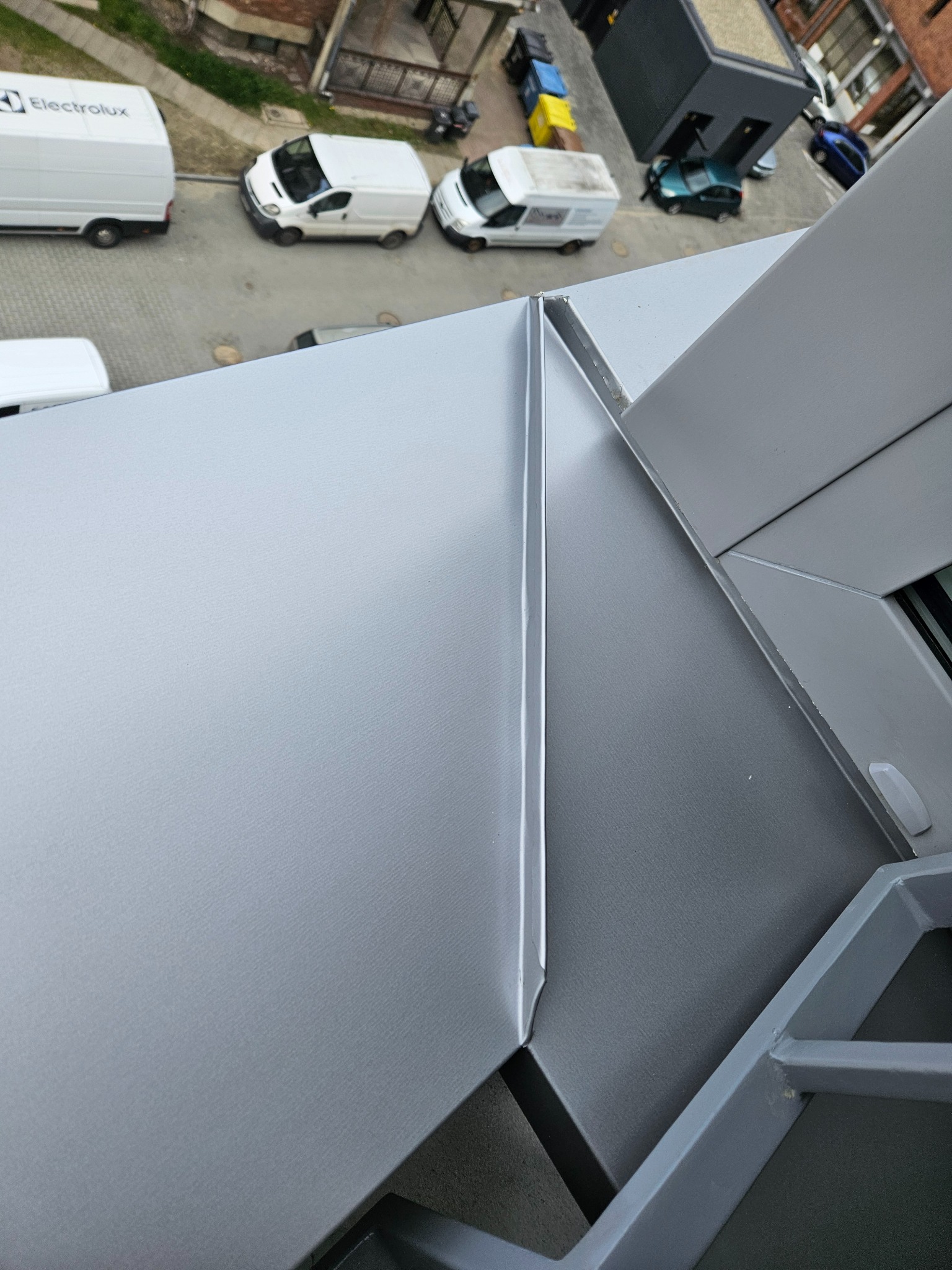 Szara, metalowa obróbka blacharska dachu z widocznym fragmentem okna i perspektywą na ulicę z zaparkowanymi samochodami dostawczymi.