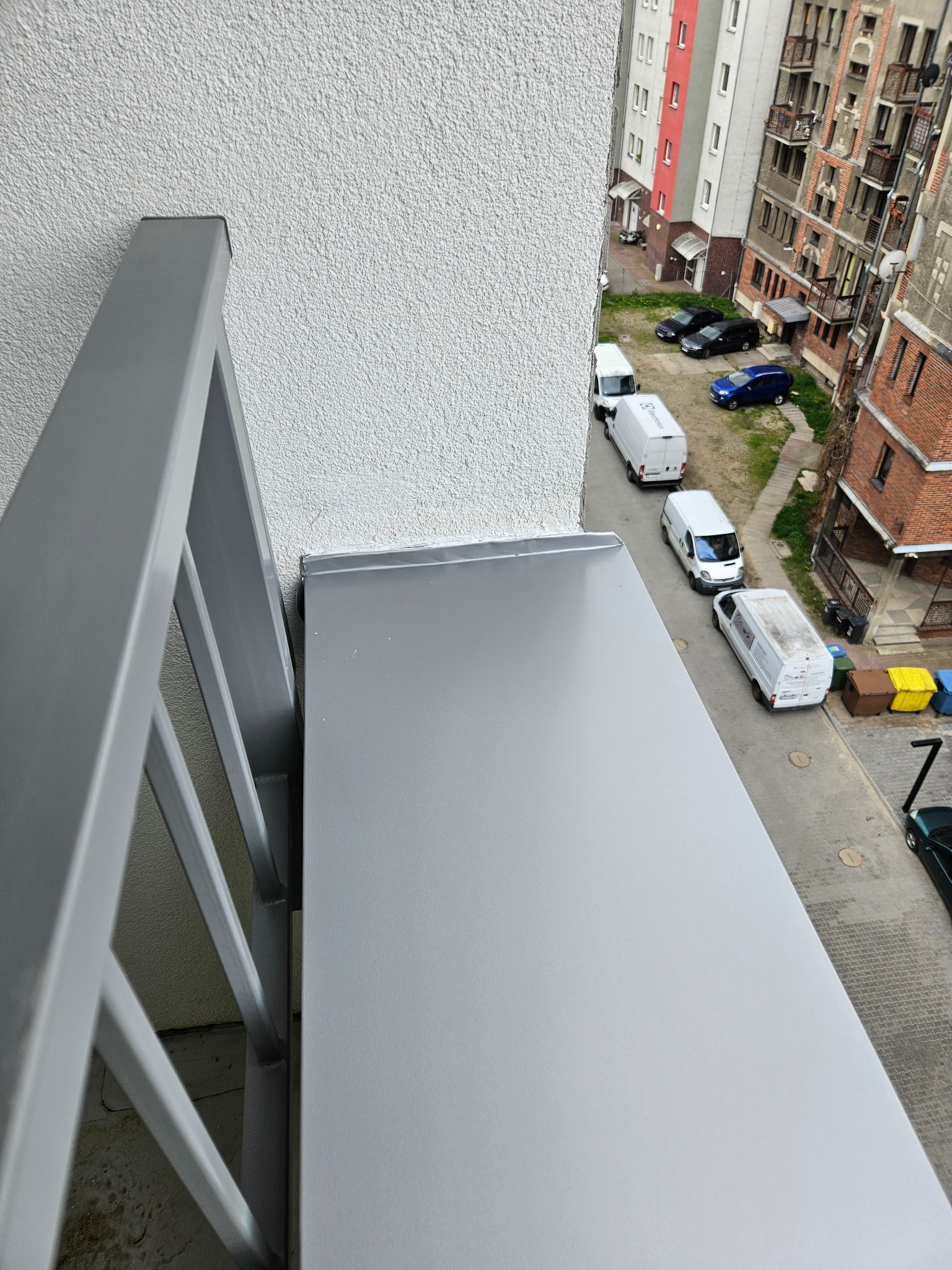 Szara, metalowa obróbka blacharska na balkonie, widok z góry na ulicę z zaparkowanymi samochodami i budynkami mieszkalnymi.