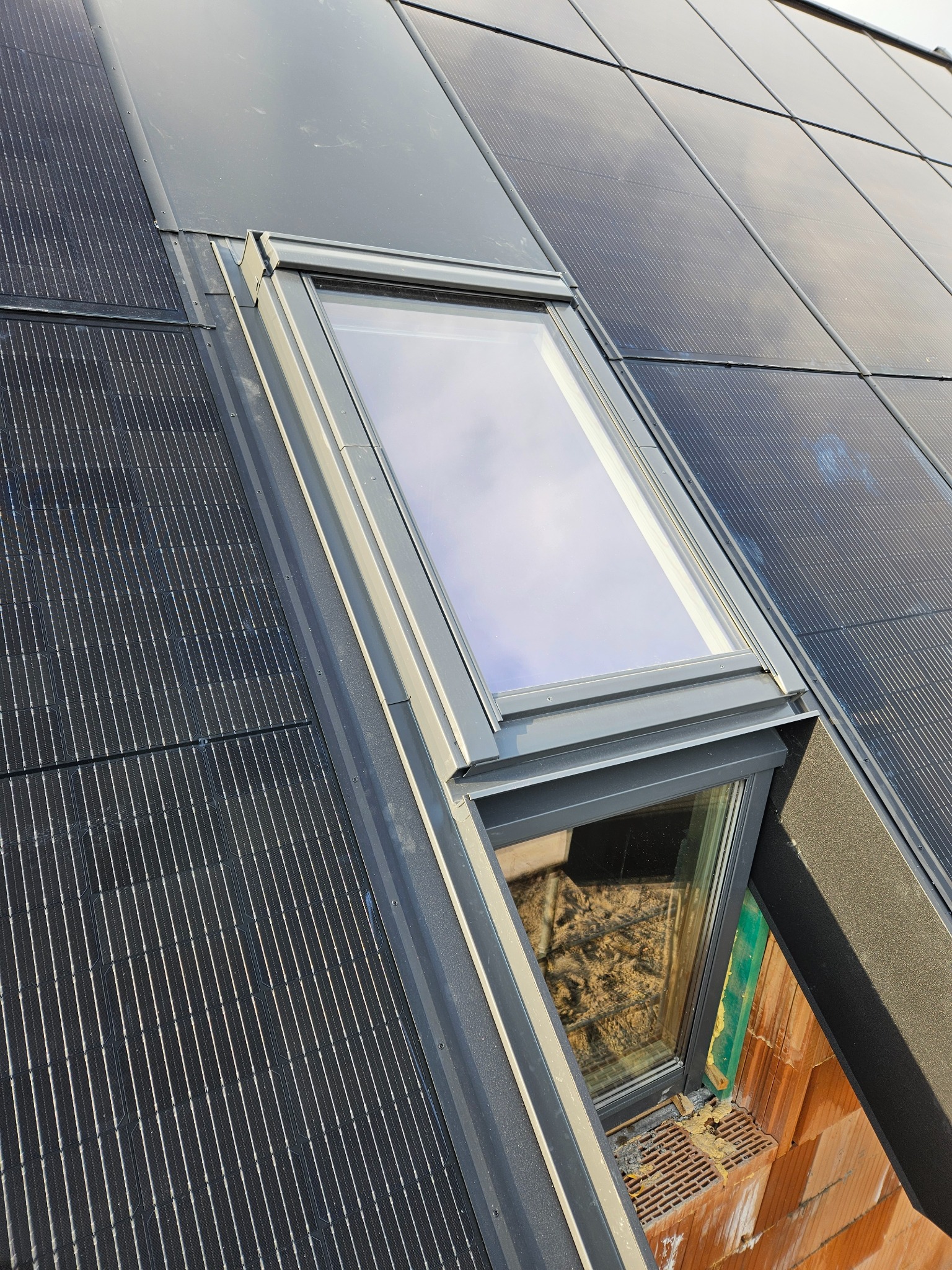 Okno dachowe z szarą ramą wkomponowane w pokrycie z paneli fotowoltaicznych na dachu, widoczne elementy izolacji i cegły wokół okna.