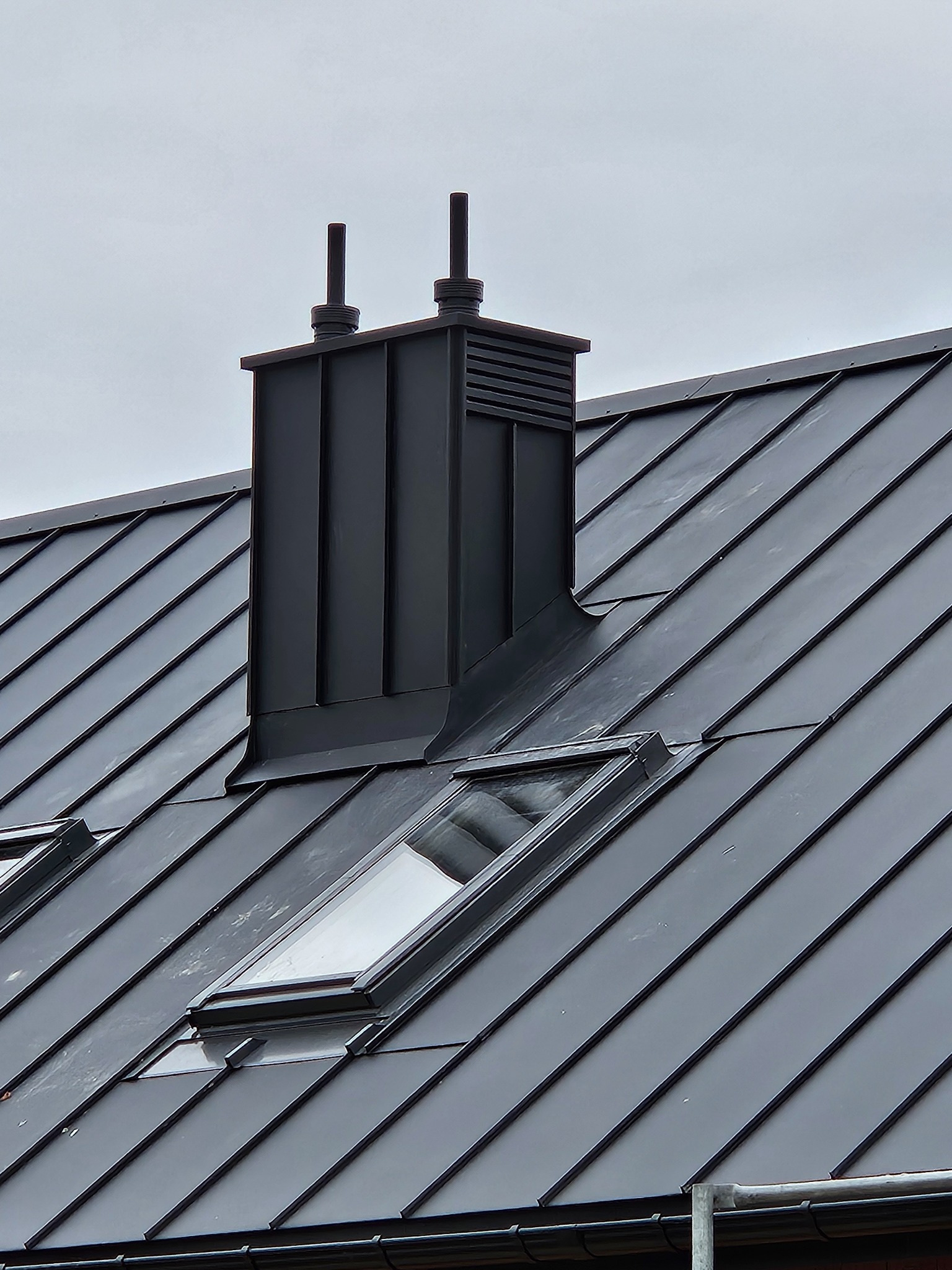 Szary dach z blachy trapezowej z oknami dachowymi i ciemnym kominem wentylacyjnym.