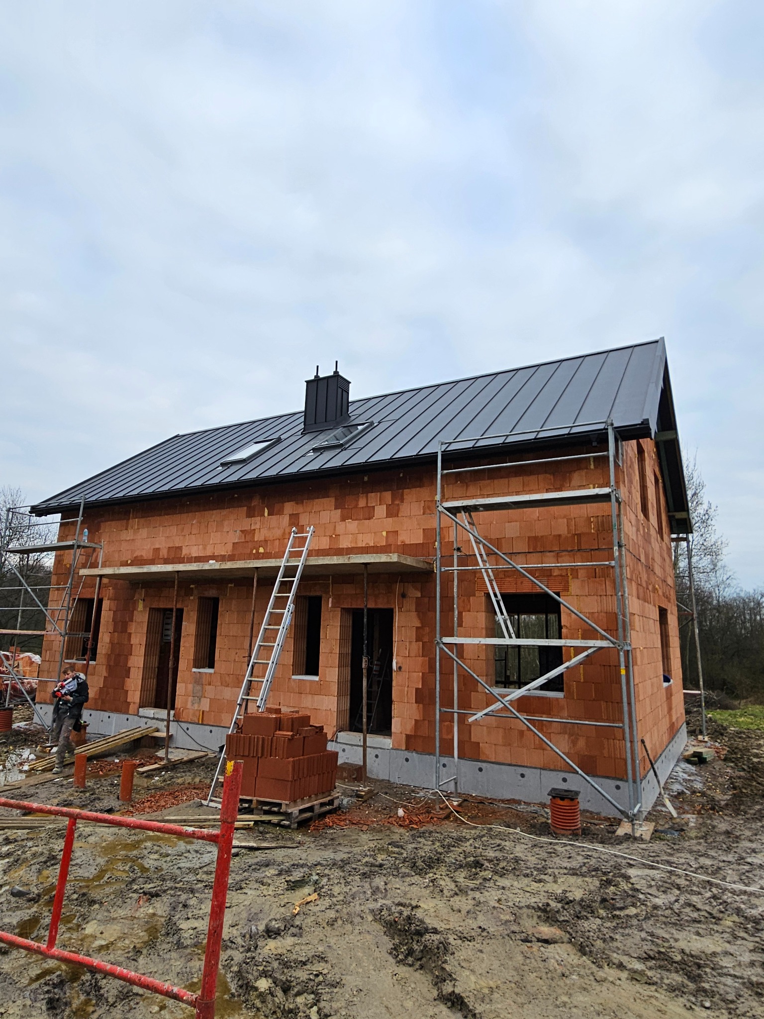 Dom w trakcie budowy z cegły, z zamontowanym czarnym dachem z blachy na rąbek stojący, rusztowania przy ścianach i stertą cegieł na palecie przed budynkiem. Widoczny pracownik w oddali.
