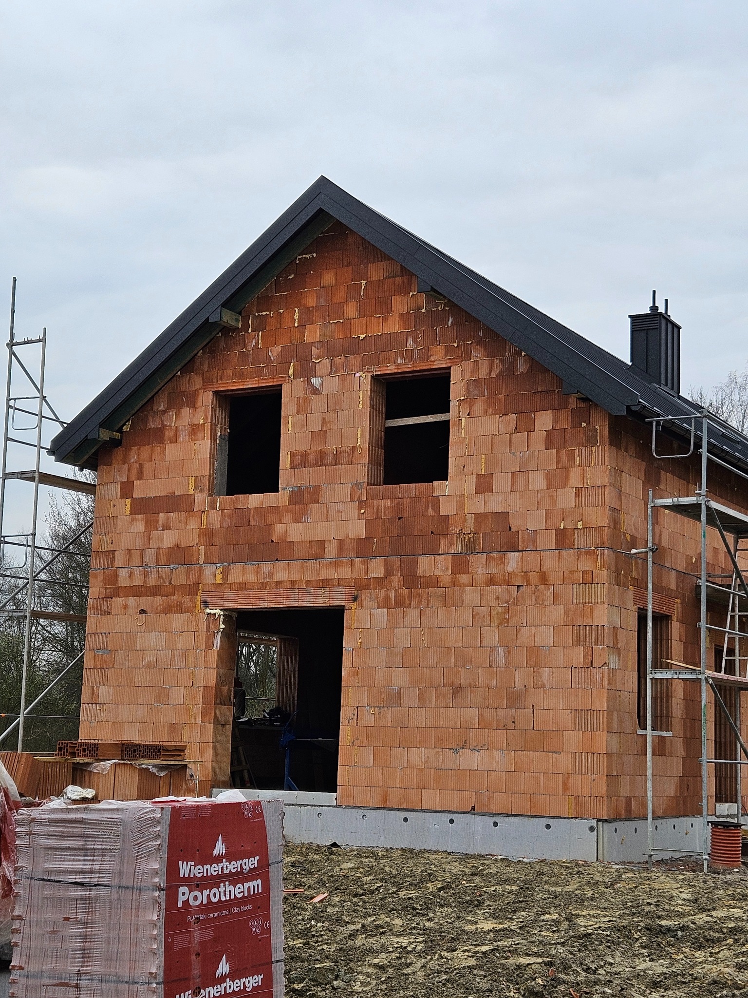 Surowy stan budynku z czerwonej cegły Porotherm, z czarnym dachem i widocznymi rusztowaniami, w trakcie budowy, z paletą cegieł na pierwszym planie.