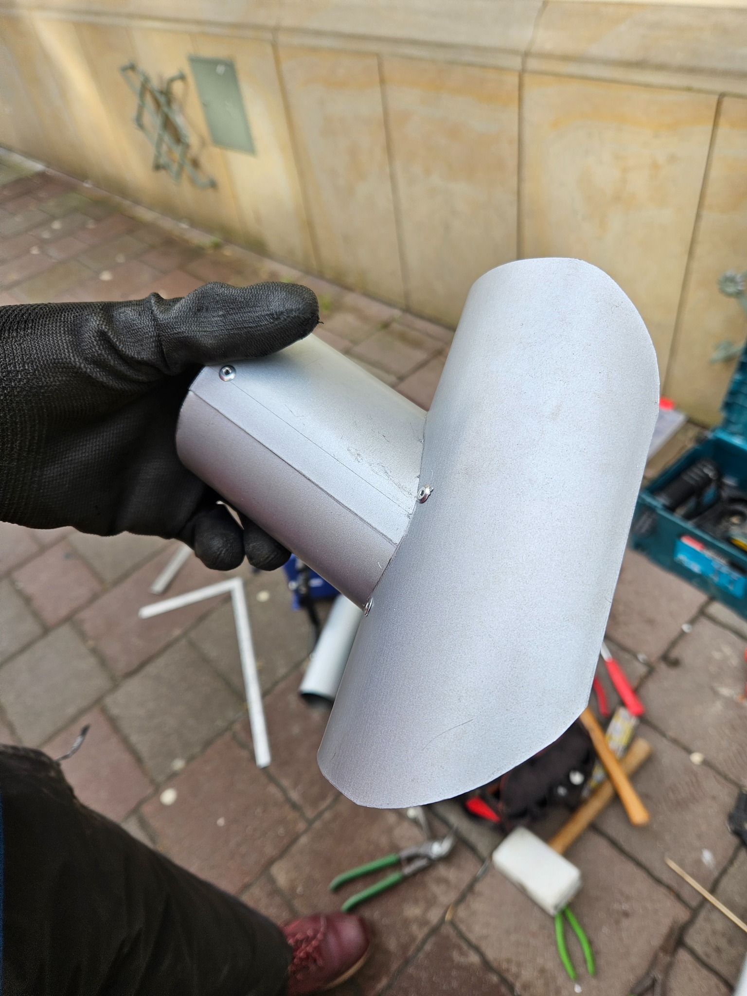 Blaszany element rynny w rękawicy roboczej, widoczne nity, narzędzia i fragment elewacji budynku w tle.