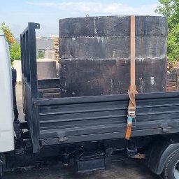 TomTransBud - Biała ciężarówka z czarną skrzynią ładunkową przewozi trzy betonowe kręgi zabezpieczone pasem transportowym na tle placu składowego z innymi elementami betonowymi.