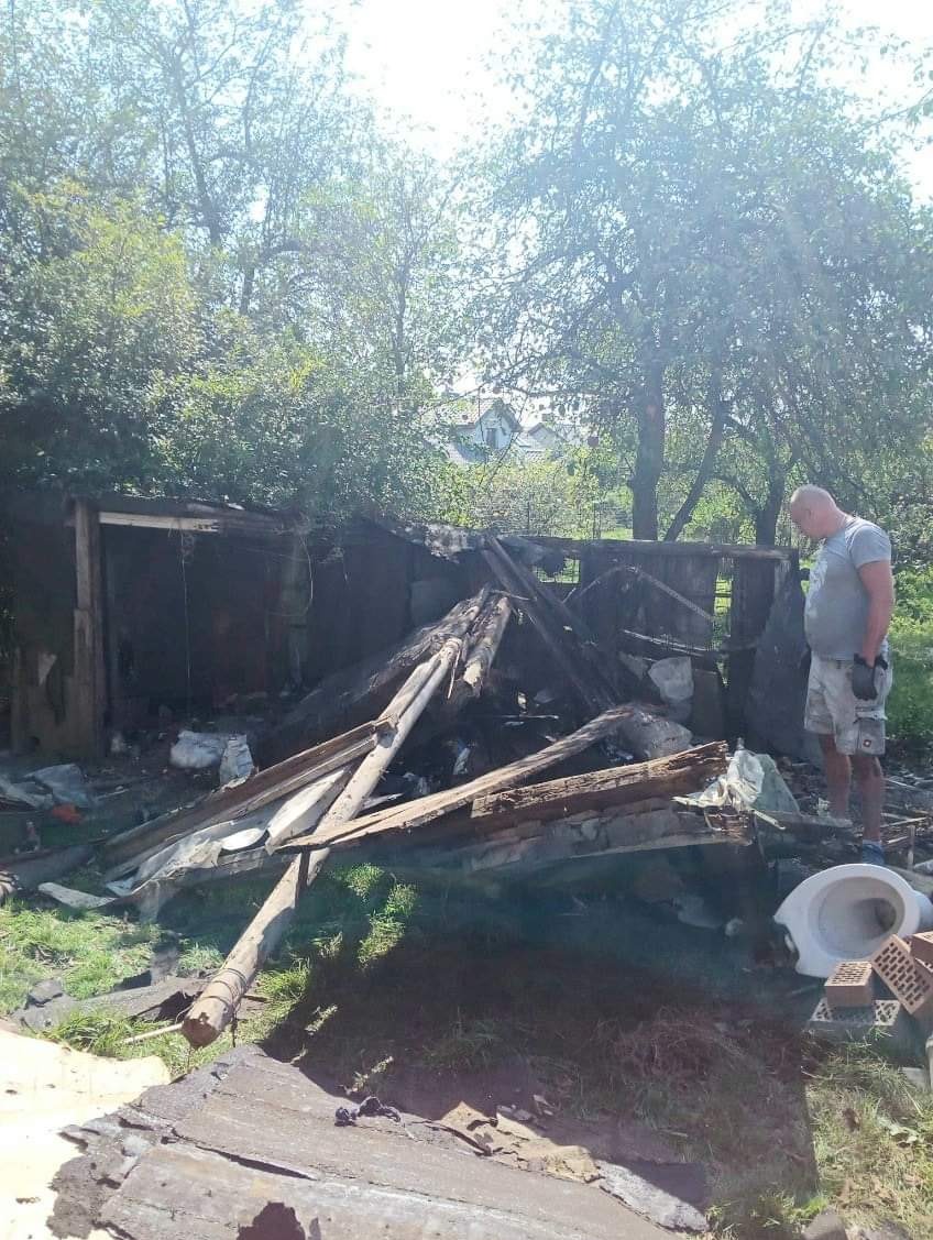 Zniszczona konstrukcja drewniana po pożarze, z stertą gruzu, cegieł i muszlą klozetową na trawie, obok stoi mężczyzna w szortach.