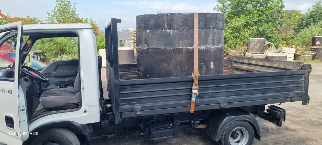 Biała ciężarówka z czarną skrzynią ładunkową przewozi trzy betonowe kręgi zabezpieczone pasem transportowym na tle placu składowego z innymi elementami betonowymi.