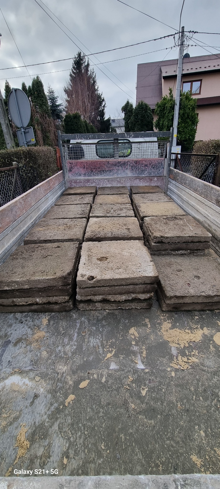 Przyczepa transportowa wypełniona starymi, betonowymi płytami chodnikowymi, widok z góry, w tle zabudowa jednorodzinna i słup elektryczny.