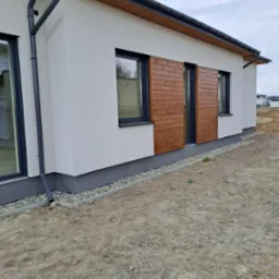 Wykończona elewacja domu jednorodzinnego z drewnianymi panelami dekoracyjnymi przy oknach, białą ścianą, szarą podmurówką i czarnymi rynnami. Teren wokół domu jeszcze niezagospodarowany.