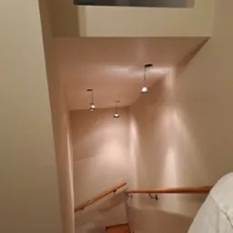 Widok z góry na klatkę schodową z drewnianymi schodami i balustradą, oświetloną trzema małymi lampami wiszącymi z sufitu, świeżo pomalowane ściany w odcieniach beżu.