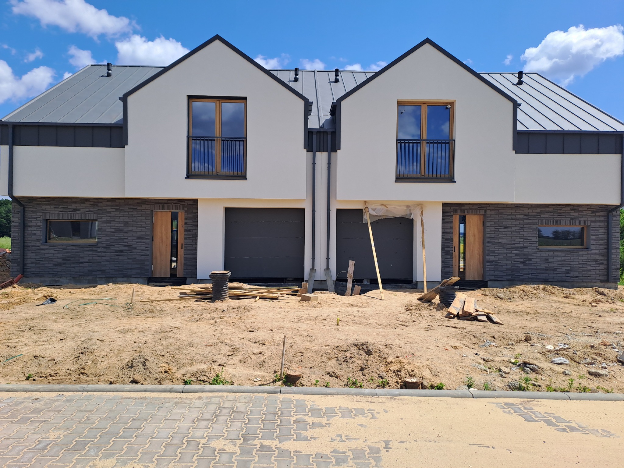 Dwa segmenty bliźniaka w trakcie budowy, widoczne szare dachy, białe ściany z elementami szarej cegły, drewniane okna z balkonami, niezadaszone garaże i teren wokół w trakcie prac ziemnych.