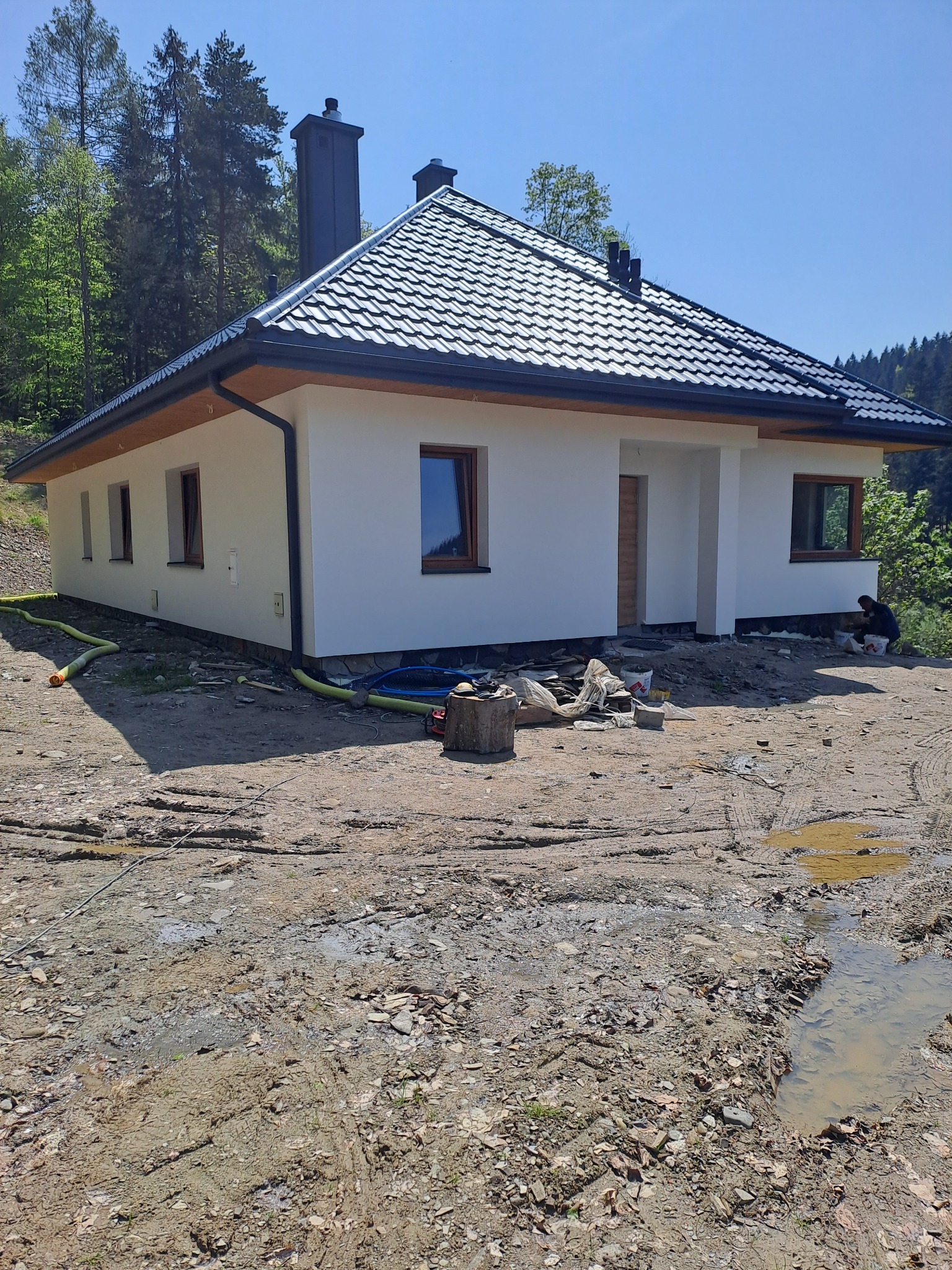 Nowoczesny, parterowy dom z ciemną dachówką i białymi ścianami po zakończeniu prac elewacyjnych, widoczne ślady prac ziemnych i przygotowania terenu wokół budynku, w tle las.