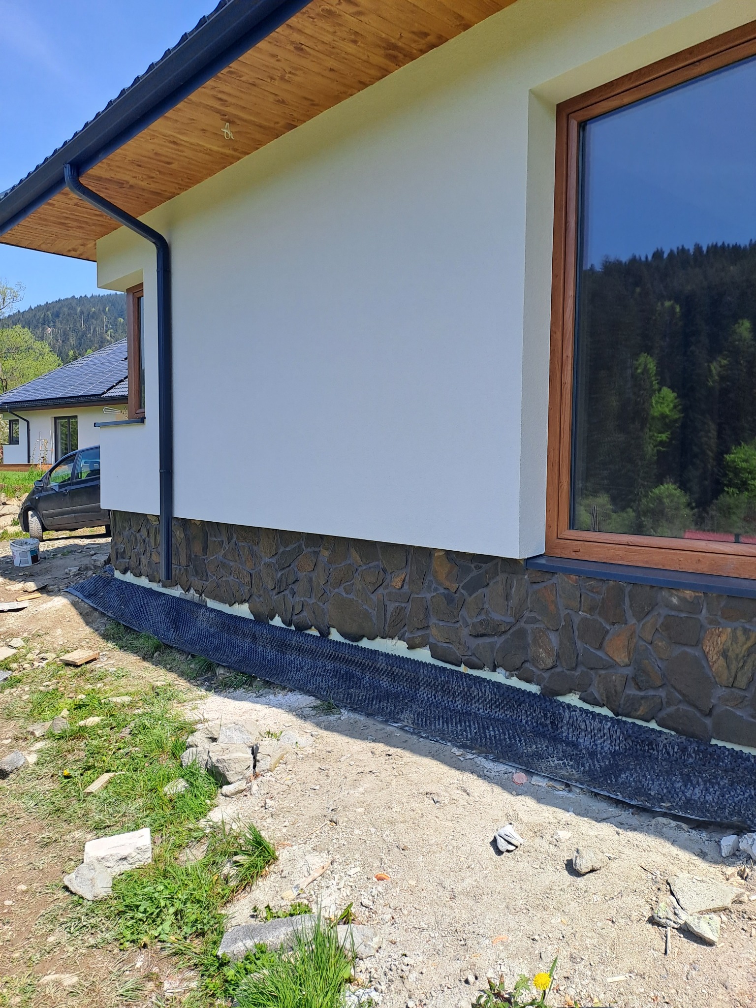 Fragment elewacji domu z jasną, gładką ścianą nad kamienną okładziną, drewnianą podbitką dachową i brązową ramą okienną, widoczne elementy systemu odprowadzania wody deszczowej i fragment...