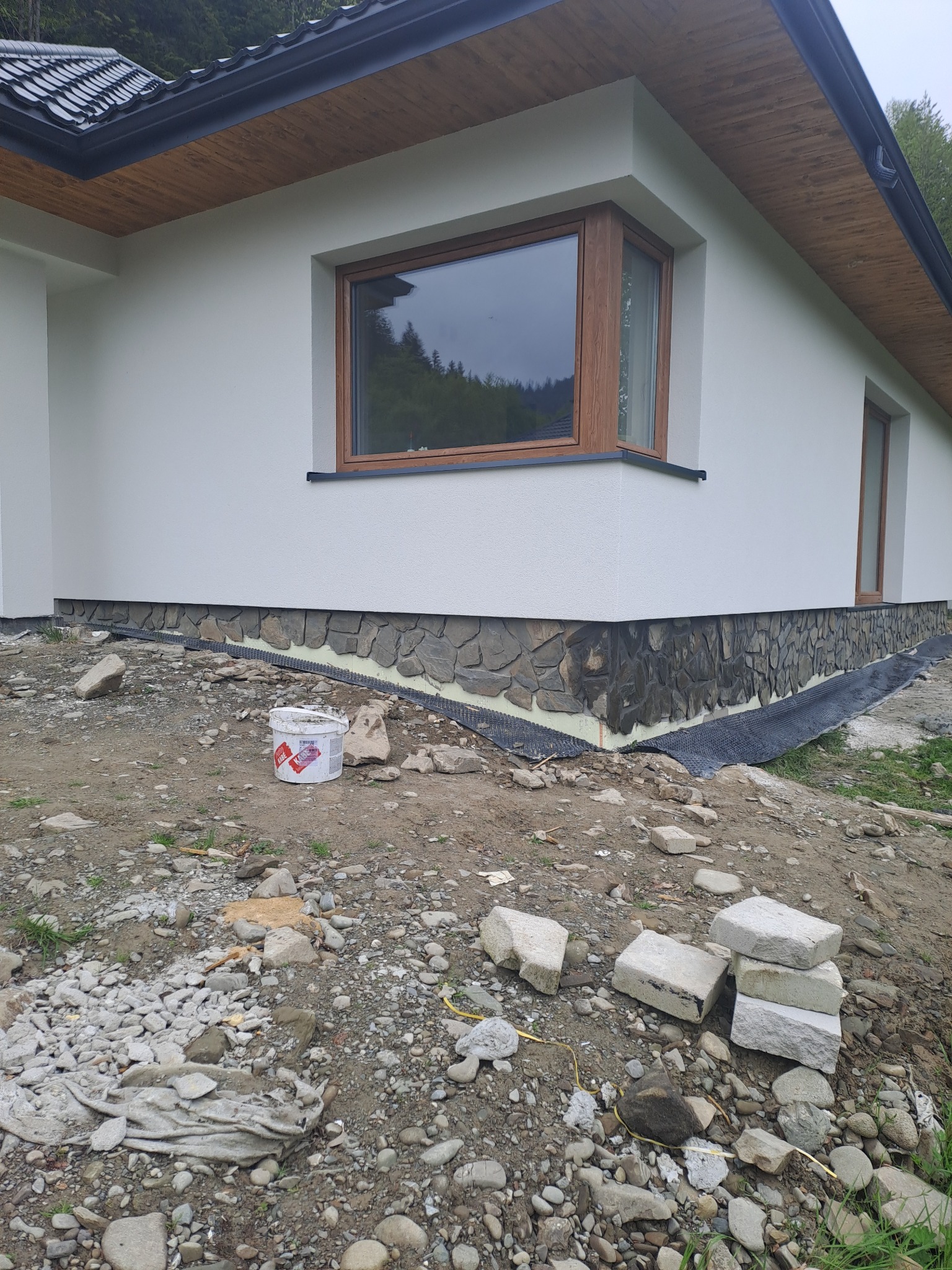 Fragment elewacji domu z oknem narożnym, widoczna warstwa izolacji i okładzina z kamienia naturalnego w trakcie montażu, kubeł farby na ziemi, teren wokół domu w trakcie prac wykończeniowych.