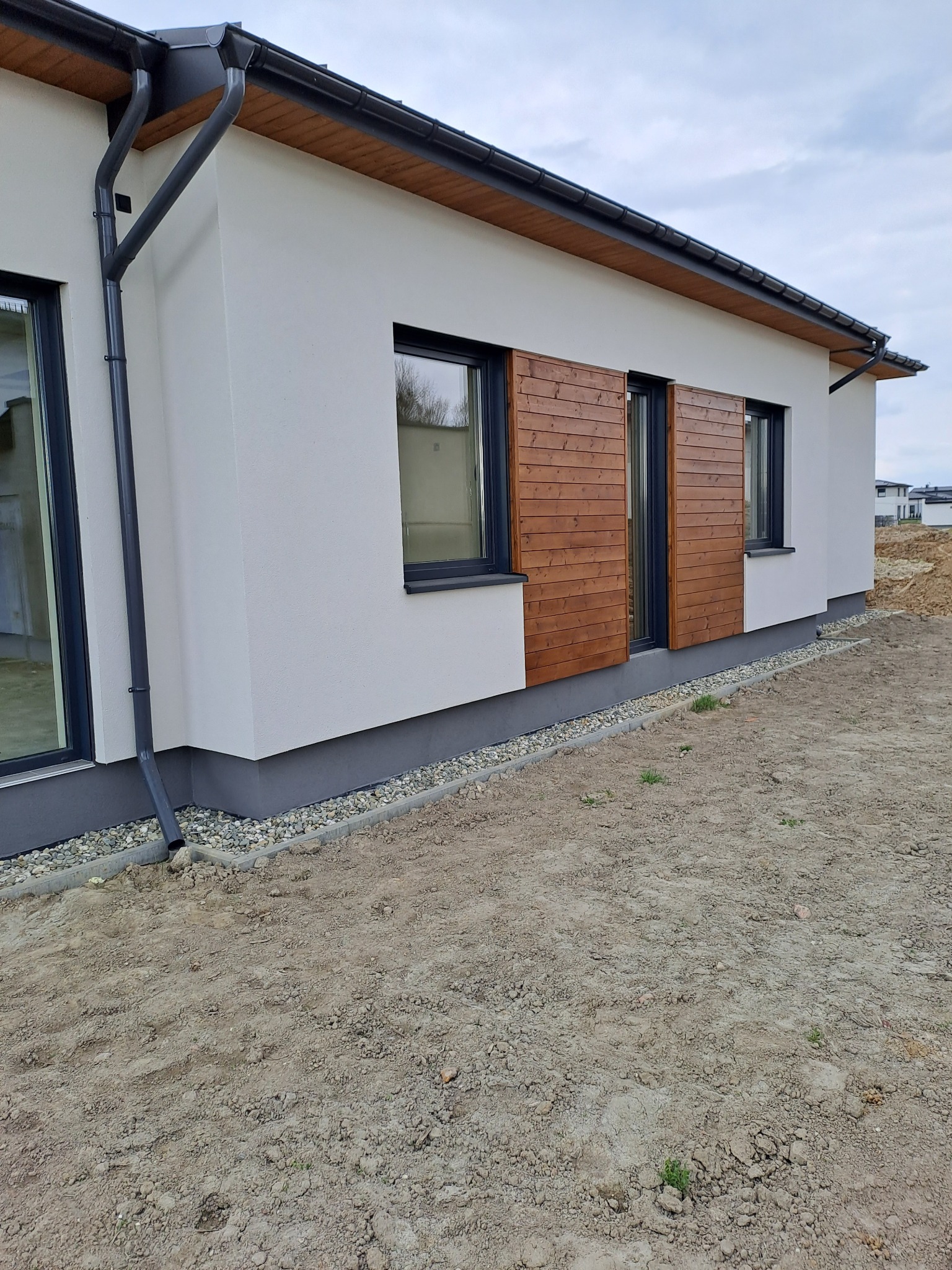 Wykończona elewacja domu jednorodzinnego z drewnianymi panelami dekoracyjnymi przy oknach, białą ścianą, szarą podmurówką i czarnymi rynnami. Teren wokół domu jeszcze niezagospodarowany.