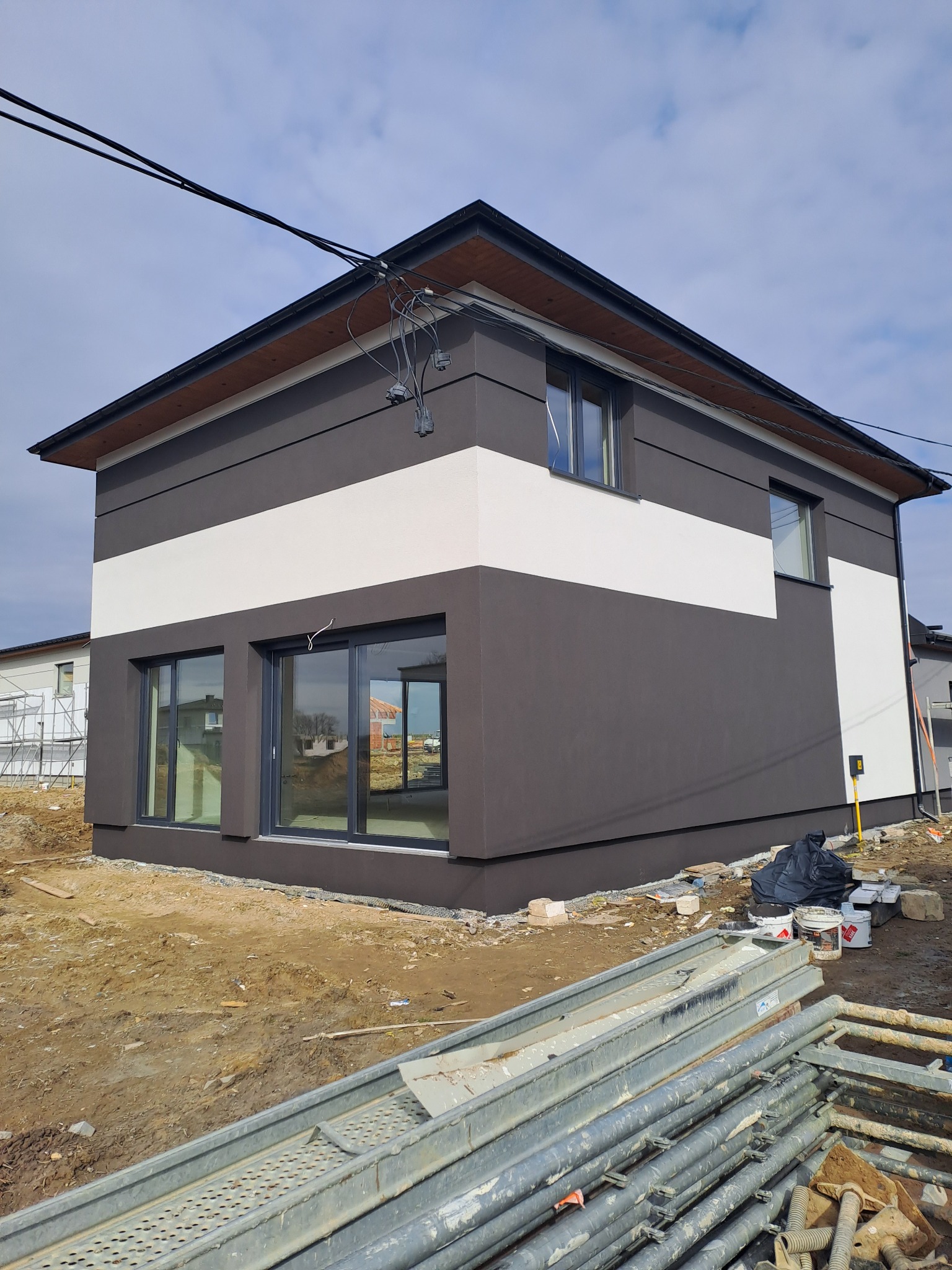 Dwukondygnacyjny dom z szarą elewacją, poziomym białym pasem i ciemną stolarką okienną, widoczne rusztowania i materiały budowlane na posesji.