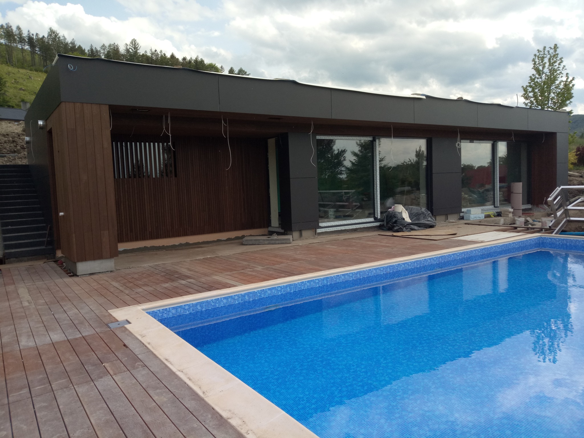 Nowoczesny budynek z drewnianą elewacją i dużymi oknami, częściowo wykończony, z zewnętrznym basenem z niebieską mozaiką i drewnianym tarasem, widoczne przewody elektryczne.