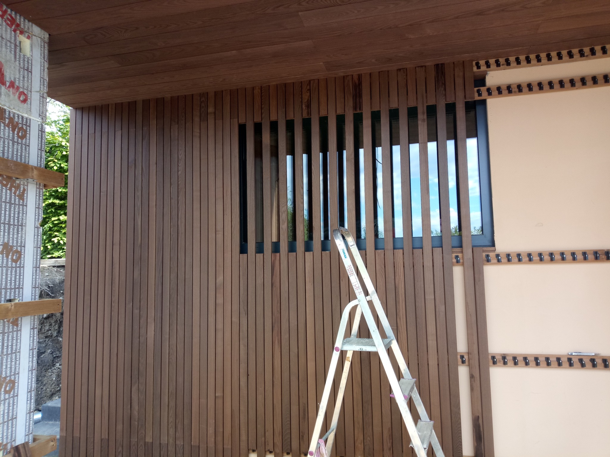 Montaż drewnianej elewacji pionowej z oknem z poziomymi lamelami, widoczna drabina i fragment ocieplenia z czarnymi uchwytami mocującymi.